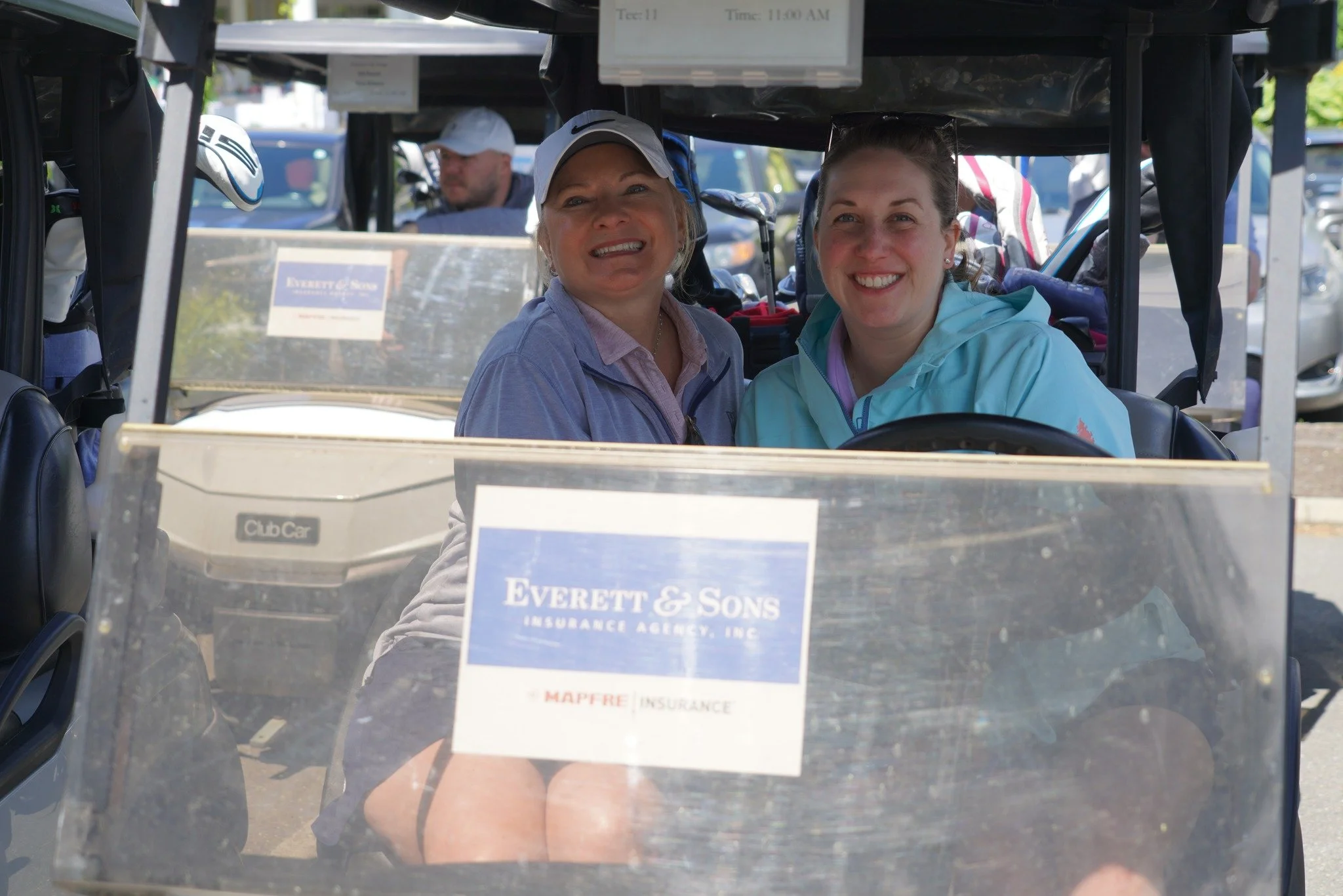 Two women sitting in a golf cart, smiling at the camera, with a man visible in the background. The golf cart has a windshield with a sign that reads 'Everett & Sons Insurance Agency, Inc.'