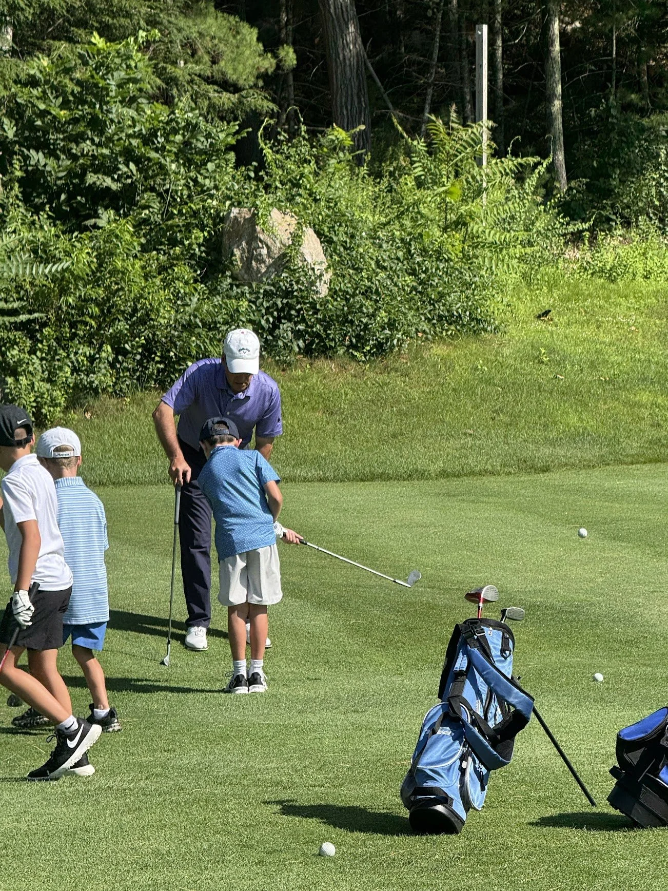 A man instructing children on a golf course, with golf bags and clubs, green grass, trees, and a bird in the background.