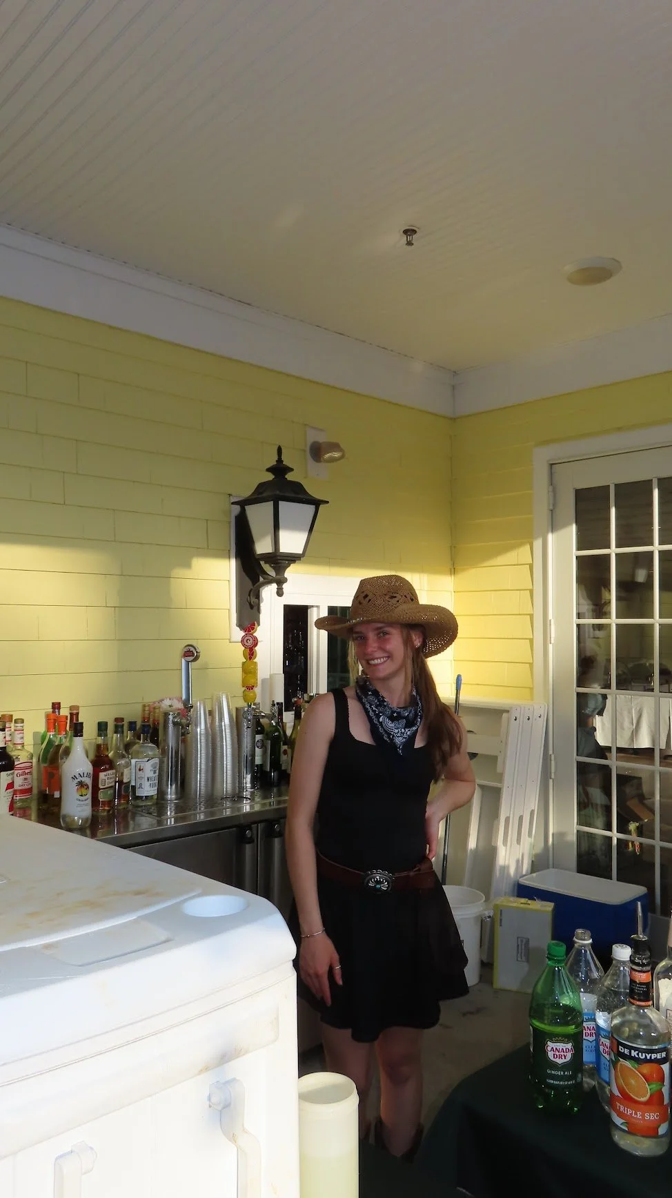 Young woman wearing a wide-brimmed straw hat and a black dress, smiling, standing behind a bar counter with bottles of alcohol and mixers, on a porch with yellow siding and a glass door.