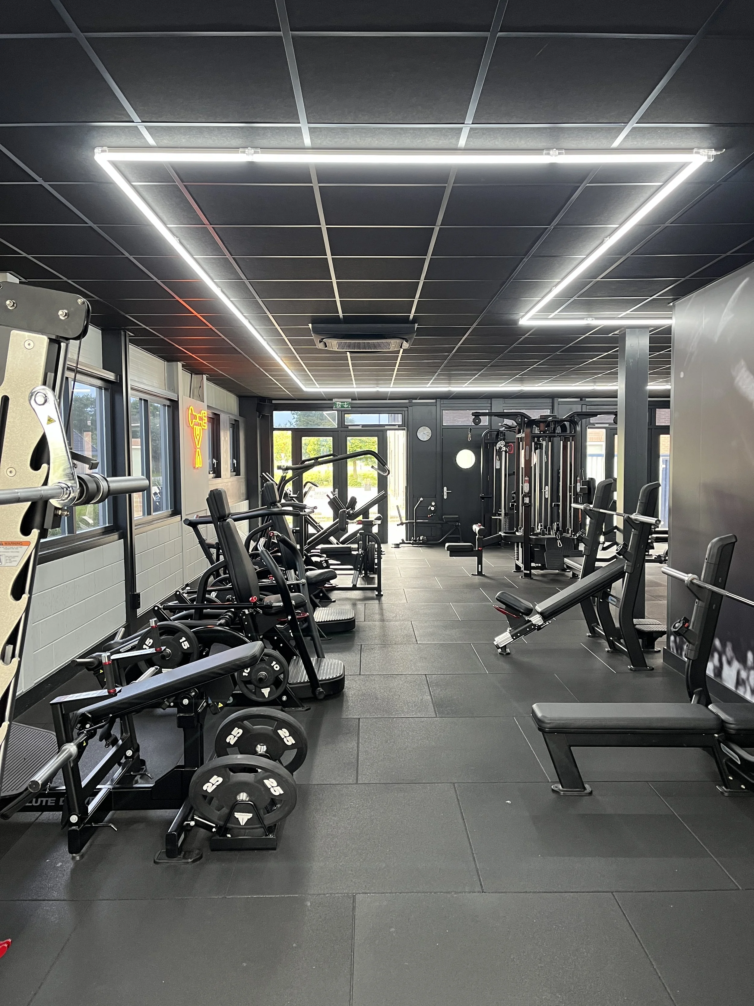 Empty modern gym with various exercise equipment including dumbbells, weight machines, benches, and cardio machines, illuminated by fluorescent lighting on a black ceiling, with windows showing trees outside.