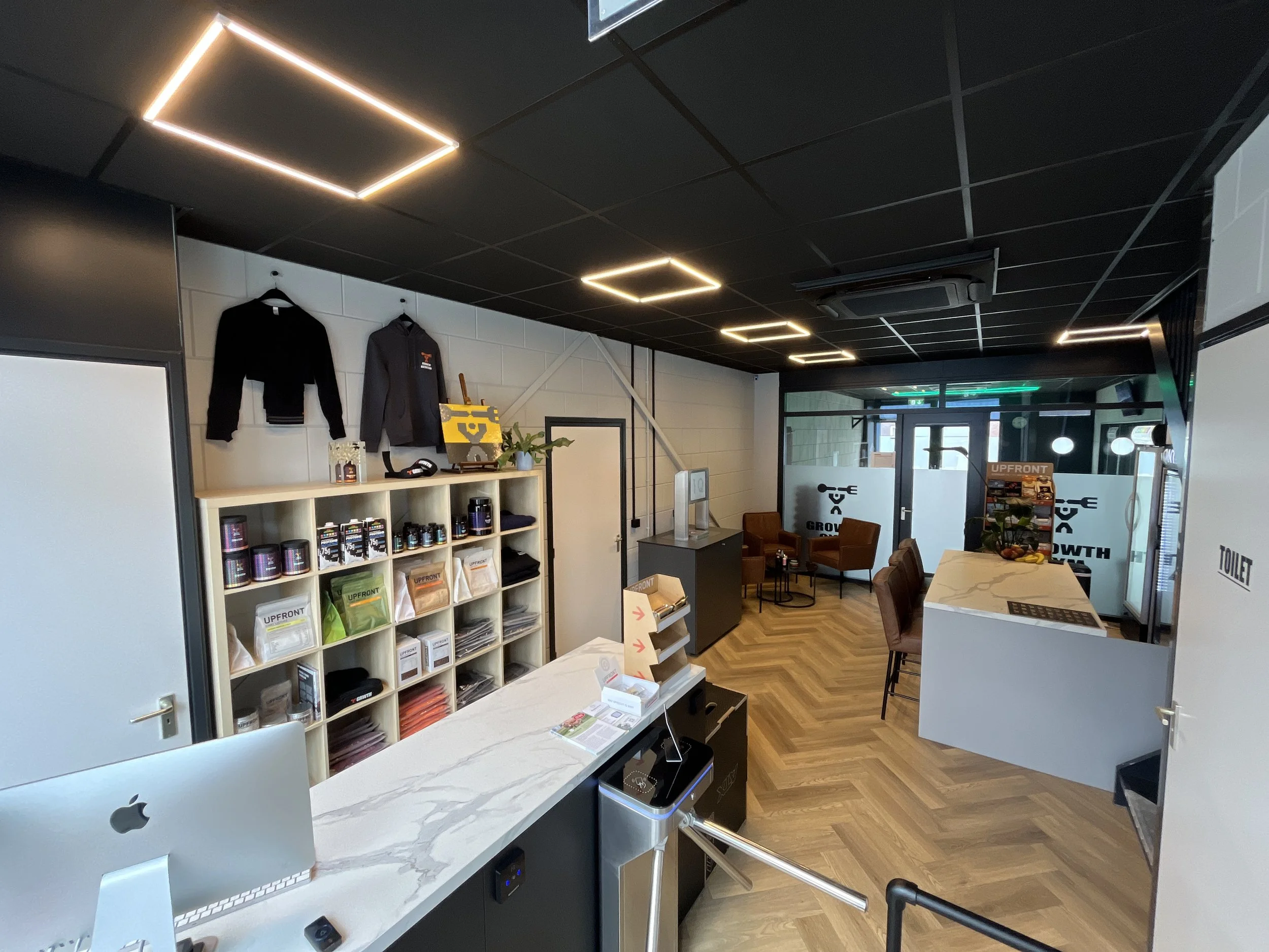 Modern reception area with a white marble counter, black ceiling with square LED lights, shelving unit with products, seating area with chairs, and a door labeled 'Toilet'.