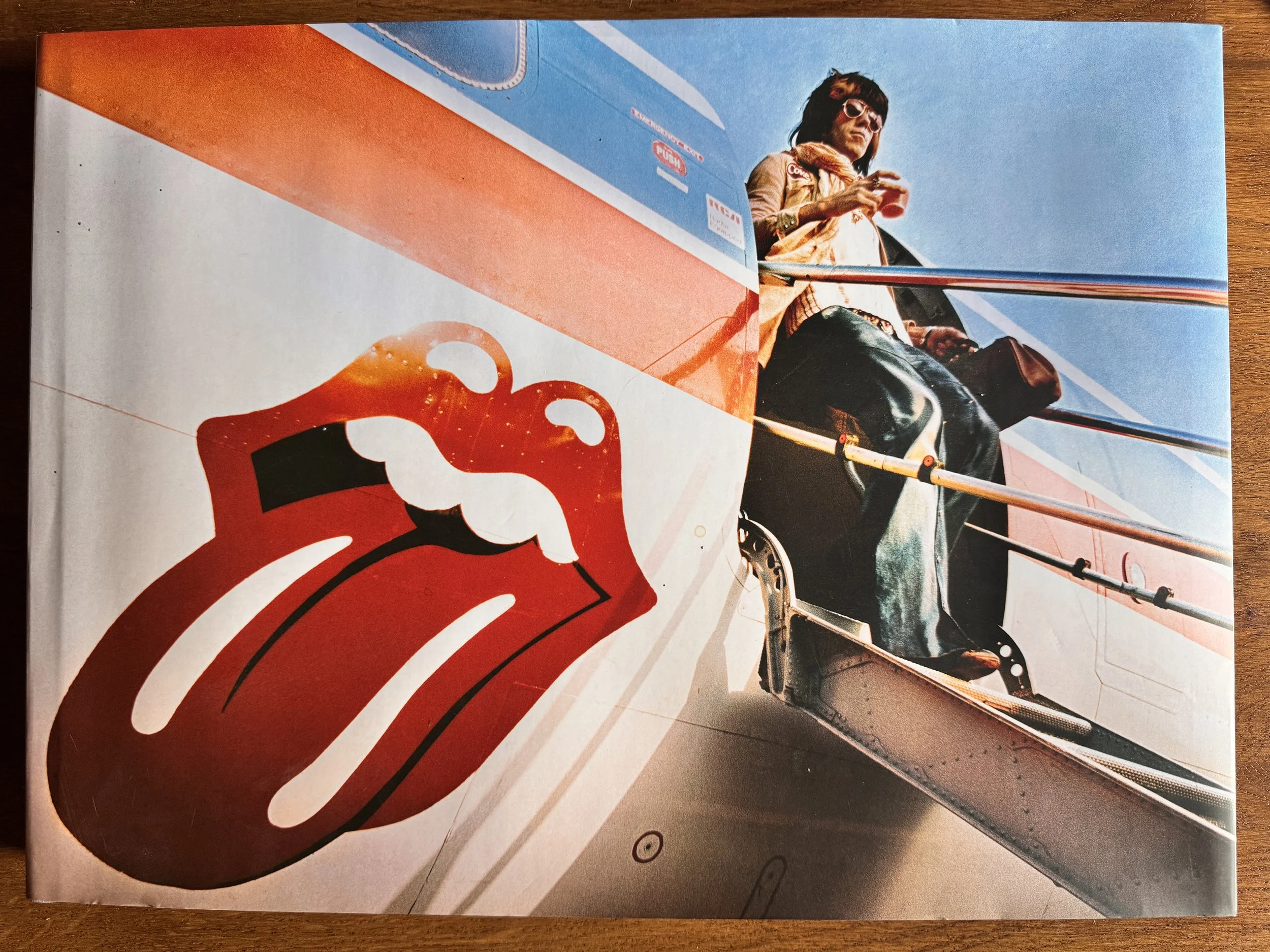 Une couverture de magazine ou un livre avec une photo d'une femme en lunettes de soleil, assise dans une voiture décapotable, tenant une tasse. La voiture est de couleur rouge et blanche et un logo rouge est visible en bas.