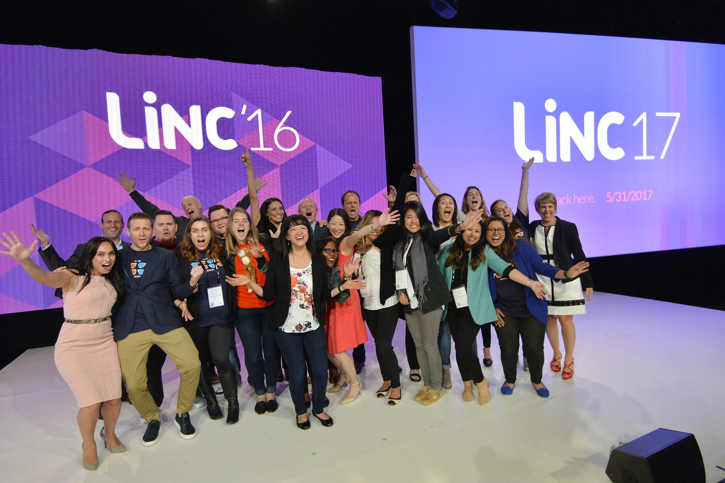 Groupe de personnes souriantes posant devant deux grands écrans affichant 'LINC '16' et 'LINC '17' lors d'un événement. 