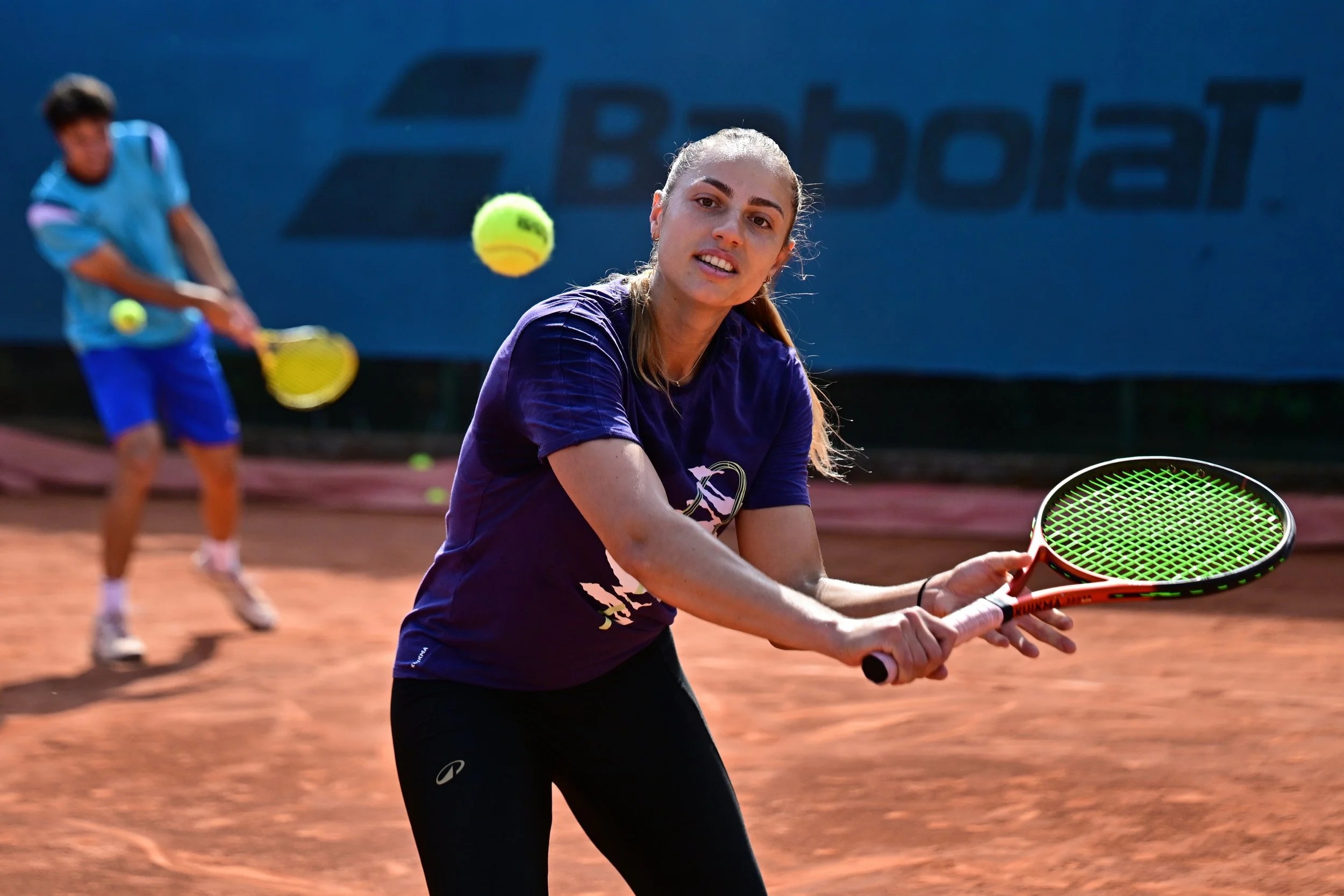 Deux personnes jouent au tennis sur un court en terre battue, femme en avant se préparant à frapper la balle, homme en arrière avec une raquette, balle en vol.