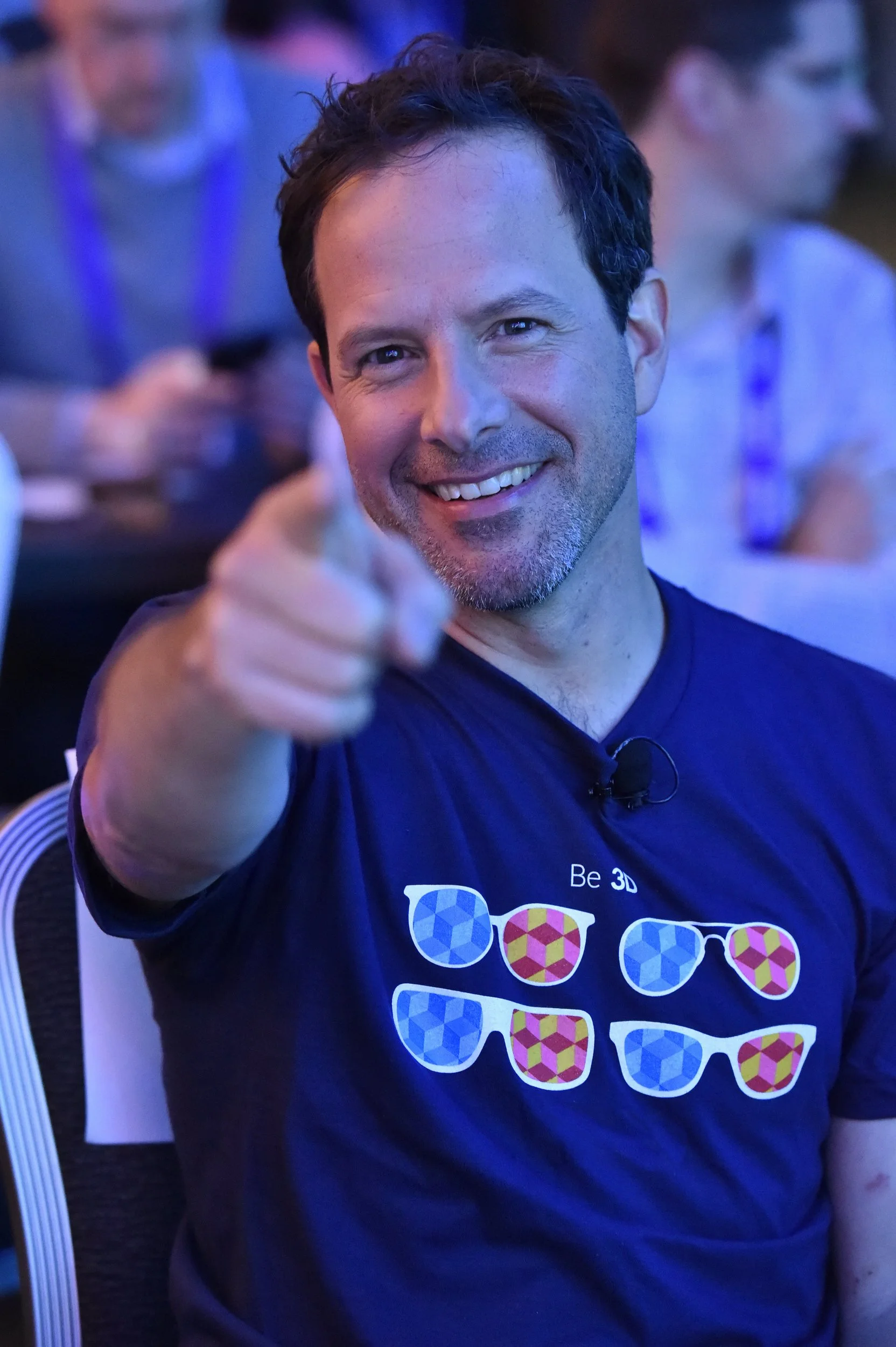 Un homme souriant pointe vers l'objectif, portant un t-shirt noir avec des lunettes colorées imprimées et le texte 'Be 3D'.