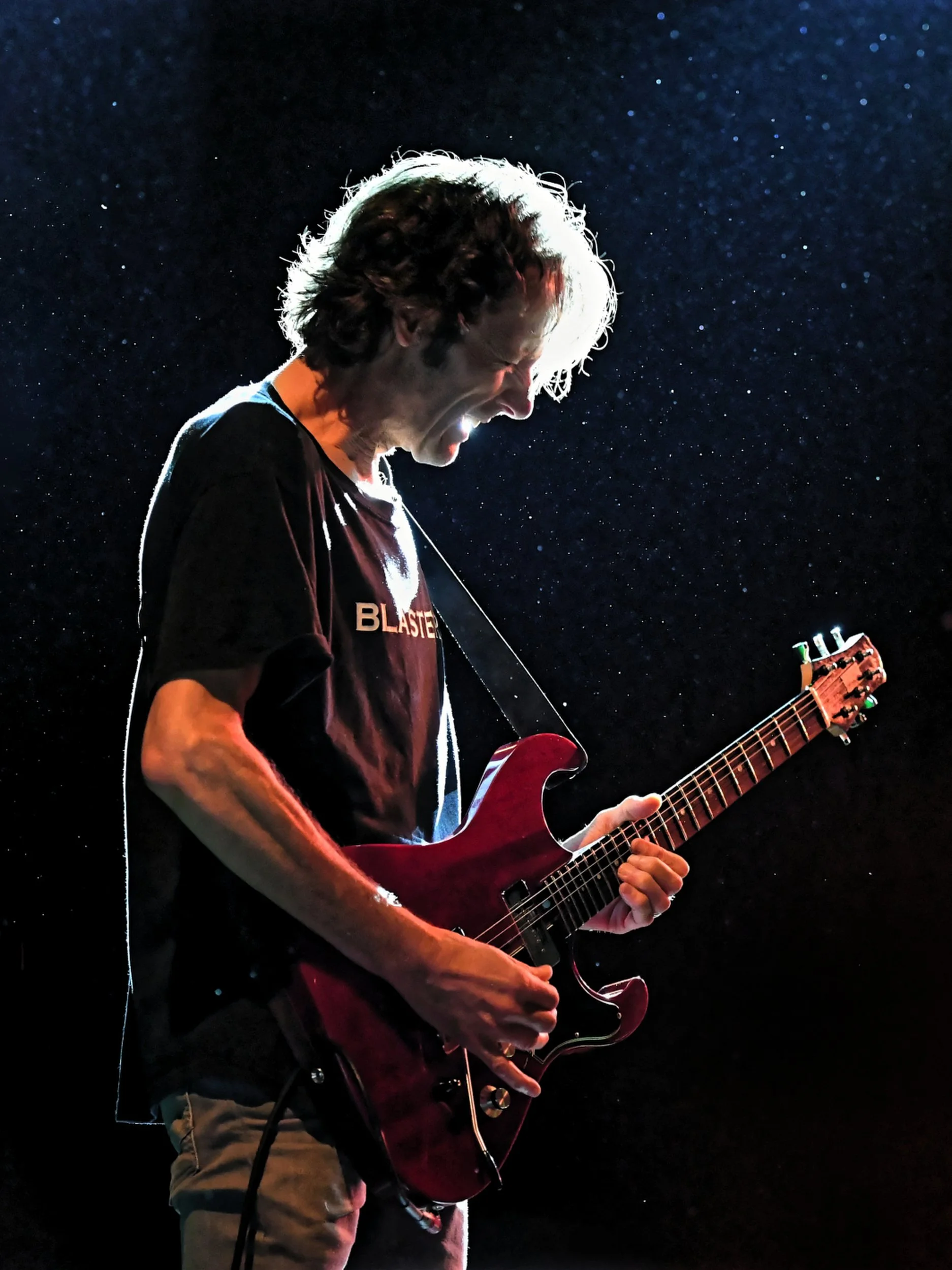 Un homme joue de la guitare électrique, portant un t-shirt noir, avec un fond étoilé.
