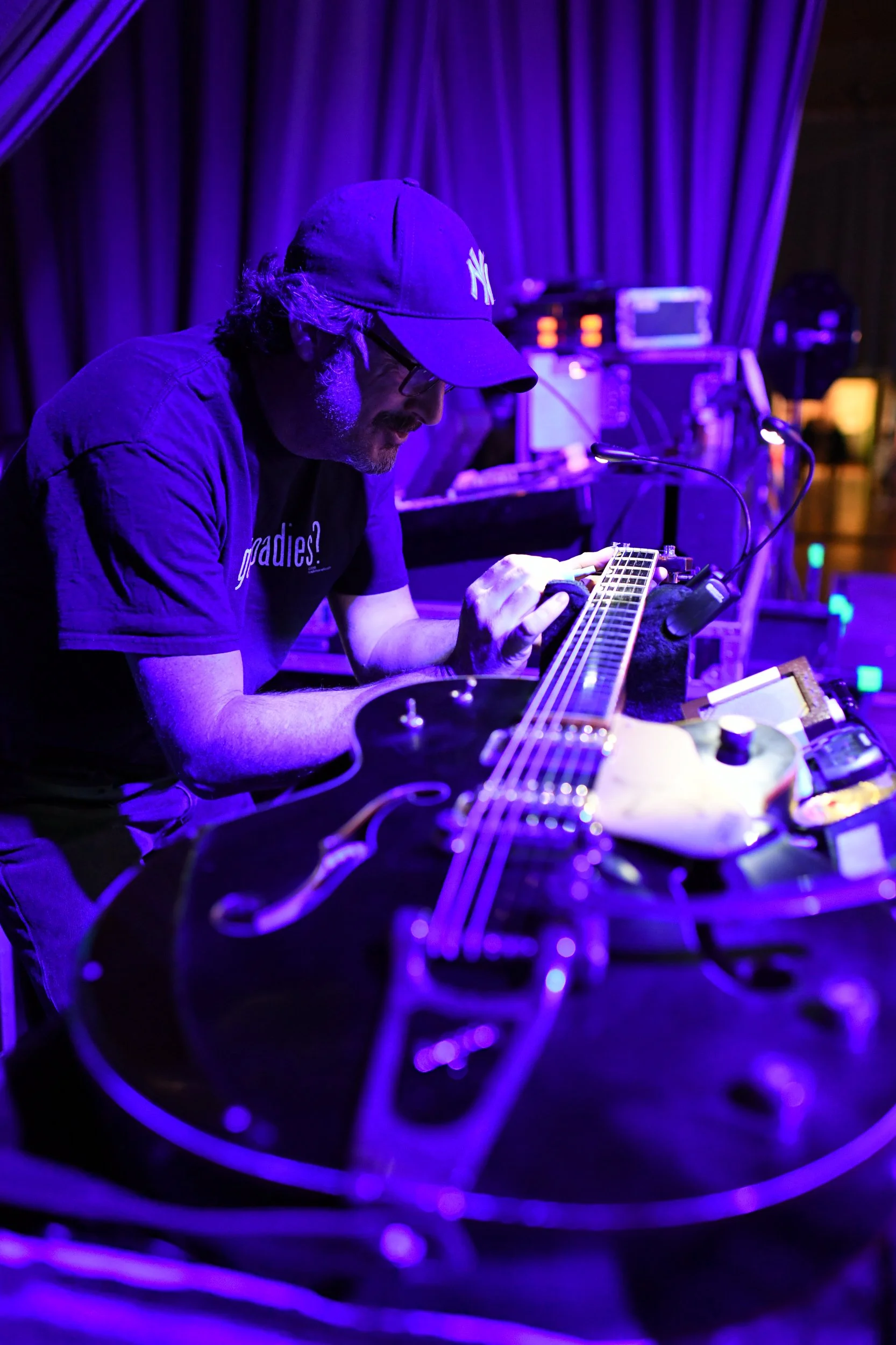Homme jouant de la guitare électrique sur scène avec des lumières violettes et un fond de rideau violet.