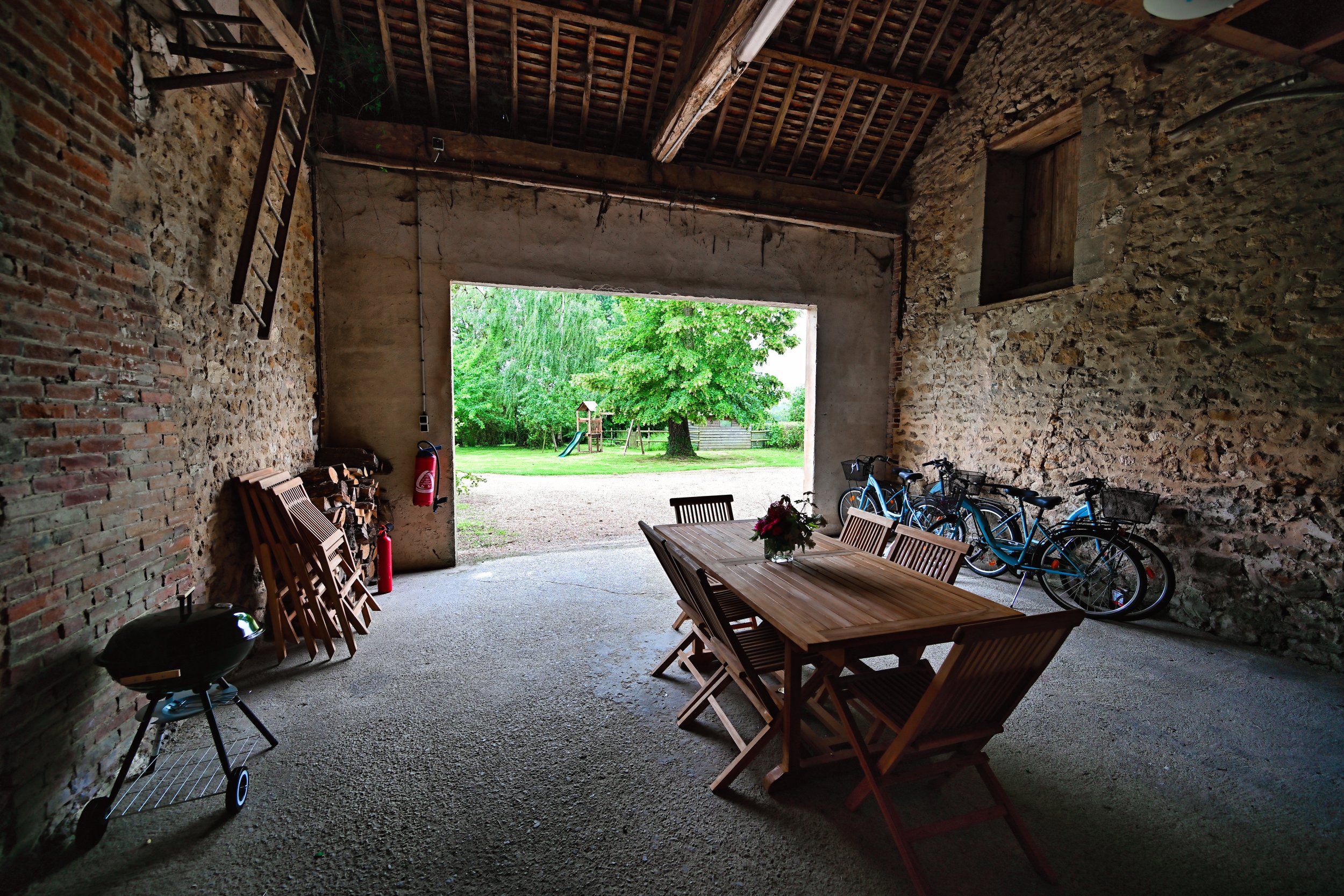 Intérieur d'une structure en pierre avec un grand ouverture donnant sur un jardin avec des arbres, une balancelle et une cabane. À l'intérieur, une table en bois entourée de chaises, plusieurs vélos et un barbecue.