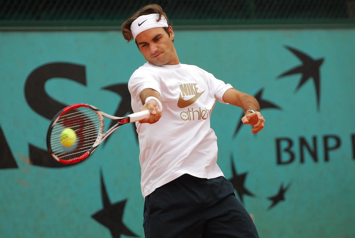 Un homme joue au tennis sur un court en intérieur, frappant une balle jaune avec une raquette. Il porte un t-shirt blanc avec un logo Nike et un bandeau blanc avec un logo Nike.