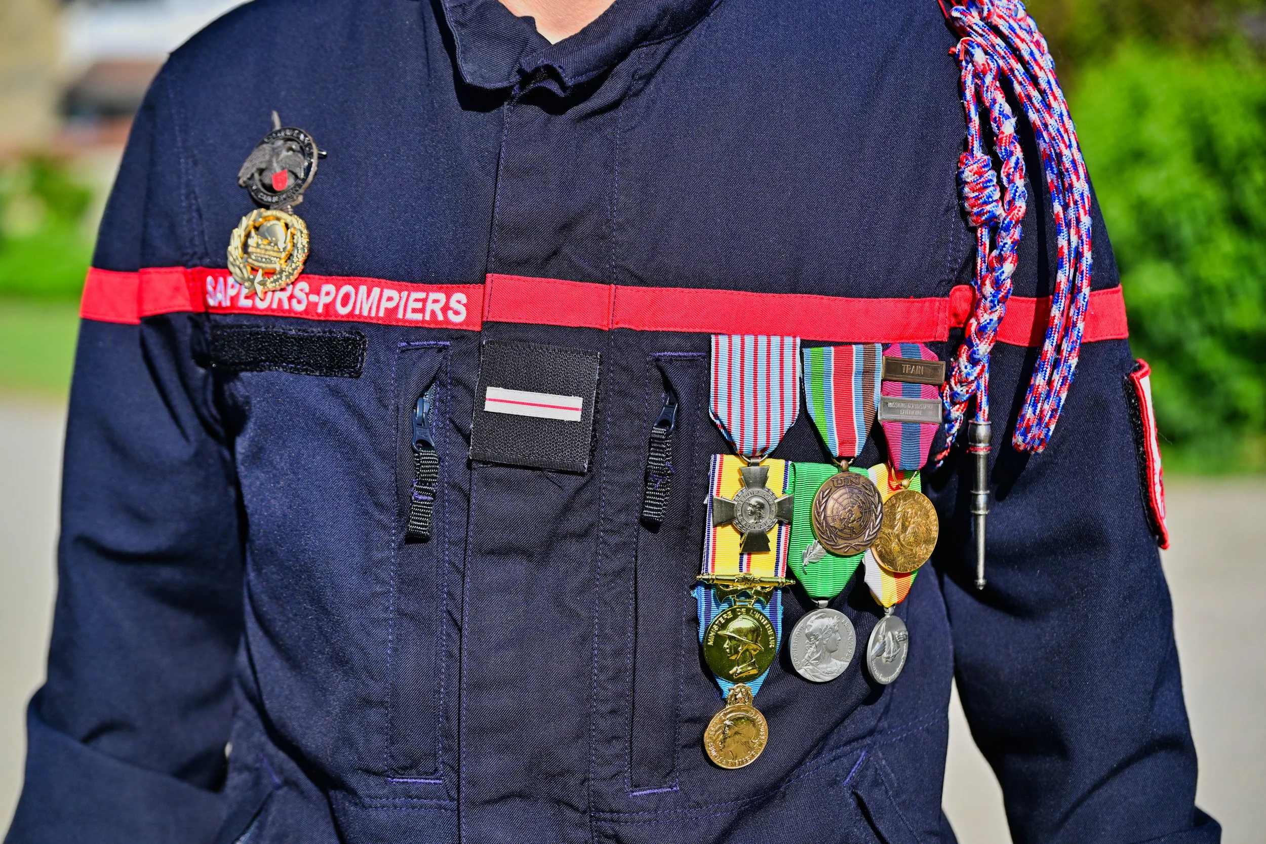 Uniforme de pompier avec médailles et ribbons, portant une écharpe bleu, rouge et blanc, dans un environnement extérieur.