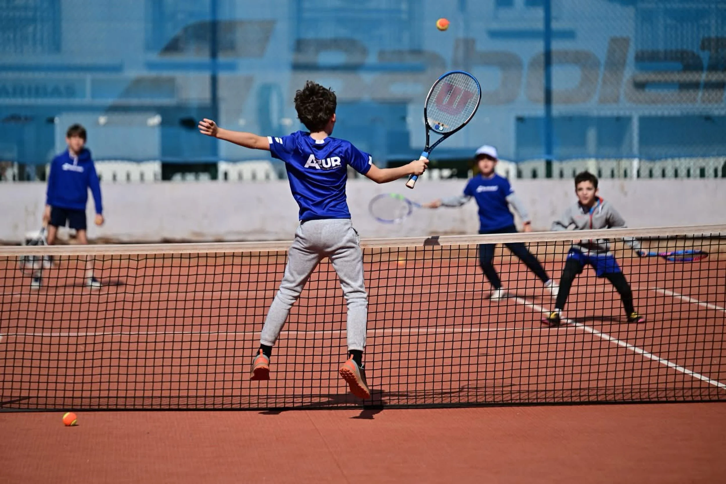 Groupe de jeunes jouant au tennis sur un court extérieur, garçon au centre prêt à frapper la balle, autres enfants en arrière-plan, filet de tennis en premier plan, bâtiment en arrière-plan.