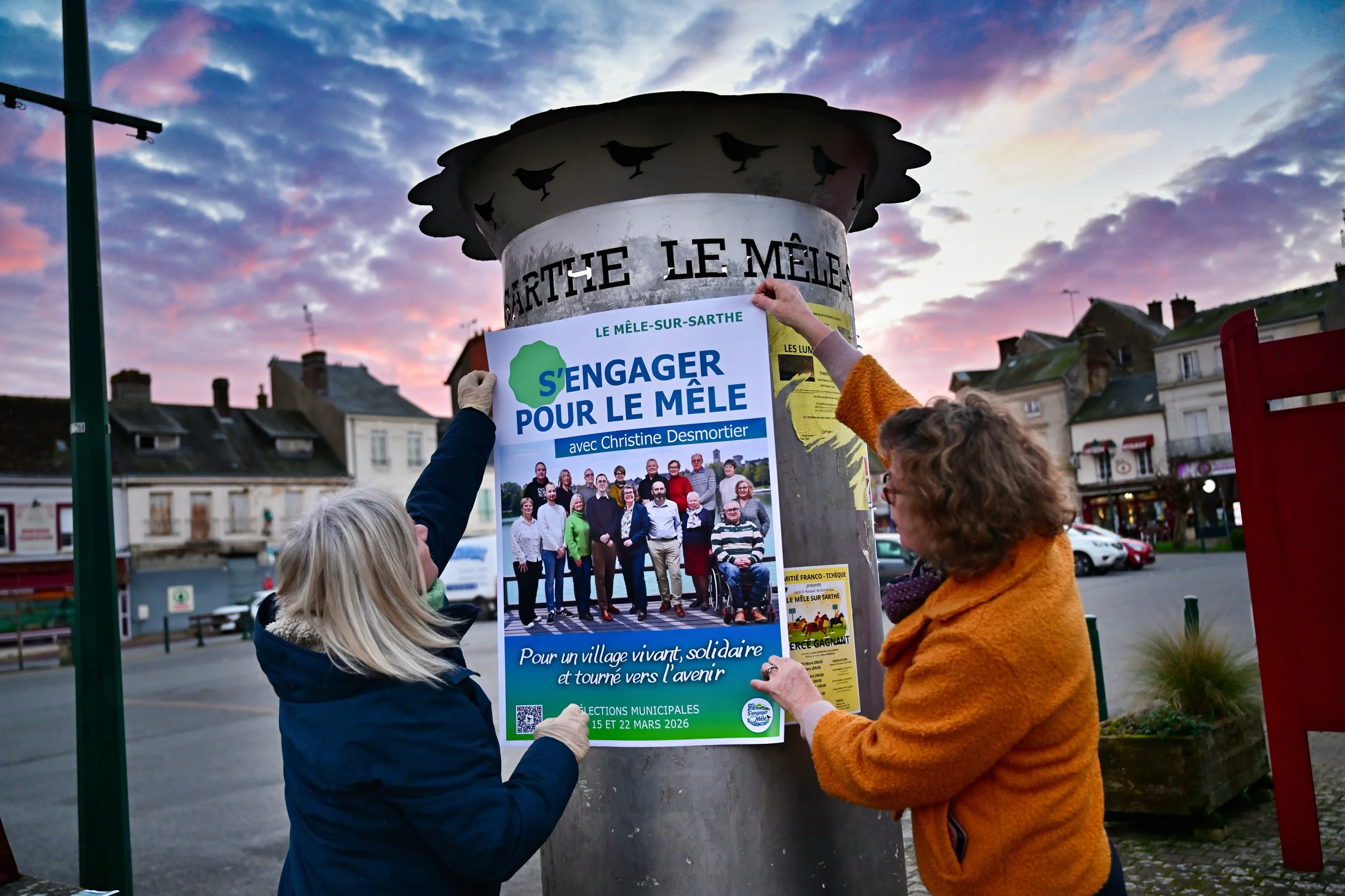 Deux femmes collent un affichage politique sur un poteau en ville lors d'un coucher de soleil. L'affiche promeut l'engagement pour le village Le Mêle-sur-Sarthe, avec une photo de groupe et des détails pour les élections municipales de mars 2026.