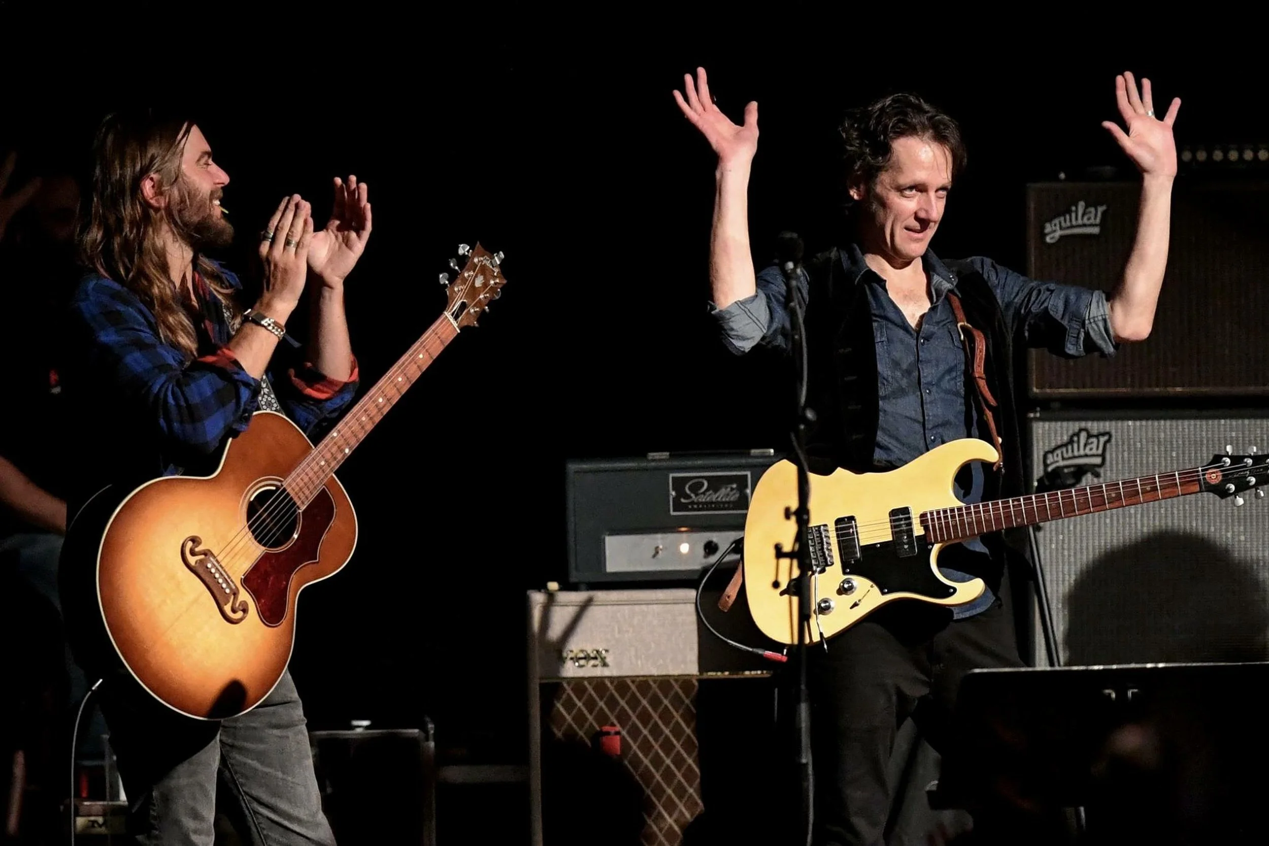 Deux musiciens jouent de la guitare lors d'une performance sur scène. Un homme a les cheveux longs et une barbe, il joue une guitare acoustique. L'autre homme, avec les cheveux courts, joue une guitare électrique. Le fond présenté avec du matériel mu