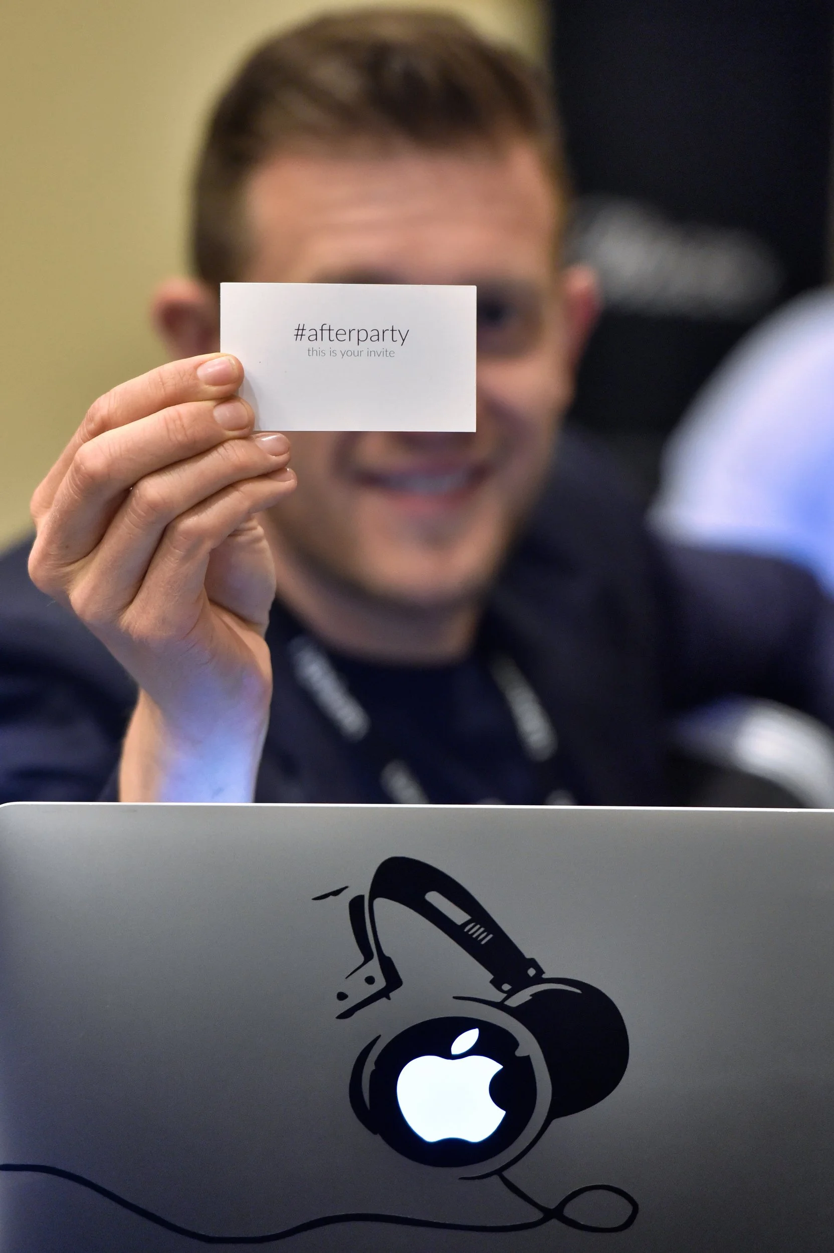 Un homme souriant tient une carte d'invitation avec le hashtag #afterparty. Une partie de son visage est floue, avec un ordinateur portable Apple visible en premier plan.