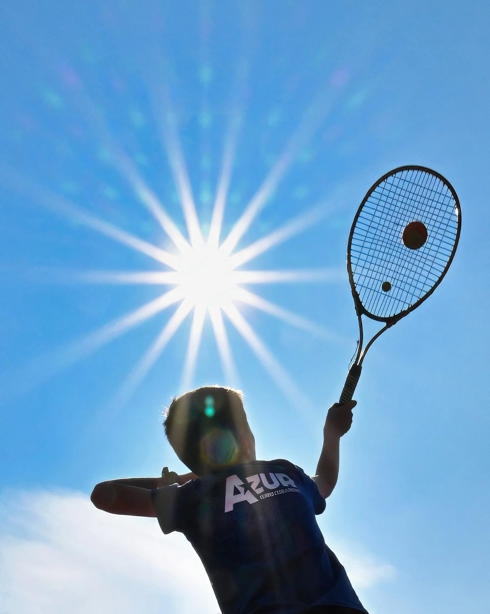 Personne jouant au tennis en plein soleil, lever de raquette vers le ciel, avec le Soleil brillant derrière, ciel bleu clair, portant un T-shirt noir.