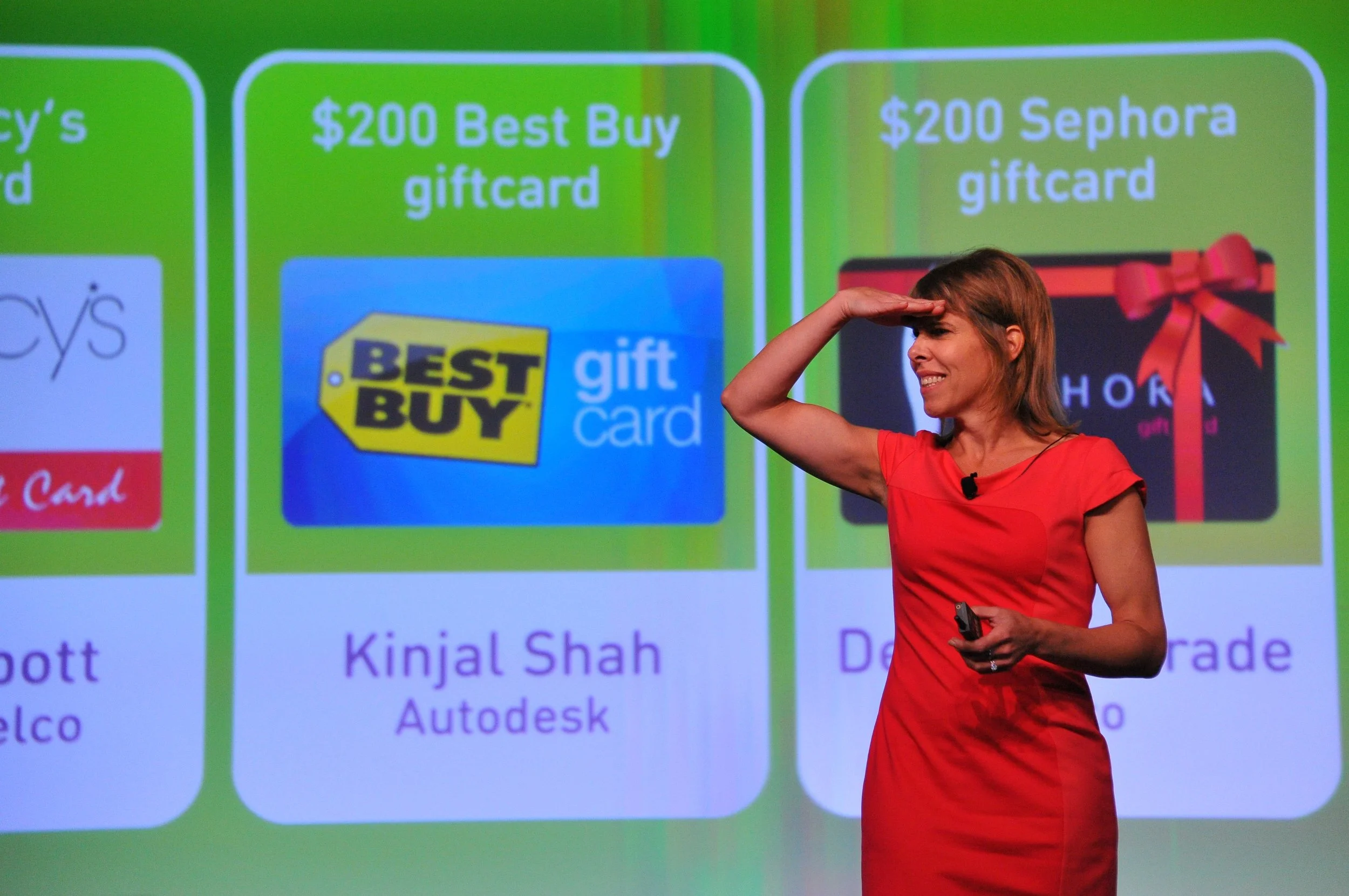Une femme en rouge sourit et fait un geste de salut à la main devant un écran affichant des cartes-cadeaux, notamment une de Best Buy et une de Sephora, d'une valeur de 200 dollars, dans un contexte de présentation ou de conférence.