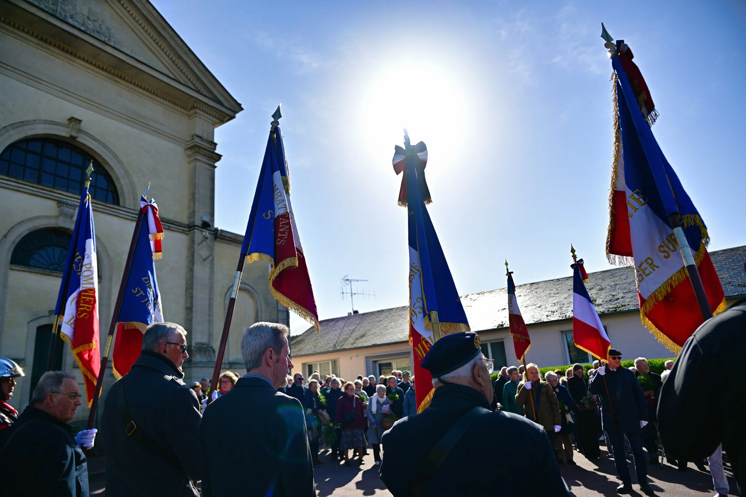 Groupe de personnes lors d'une cérémonie commémorative avec des drapeaux colorés sous un ciel ensoleillé devant un bâtiment historique.