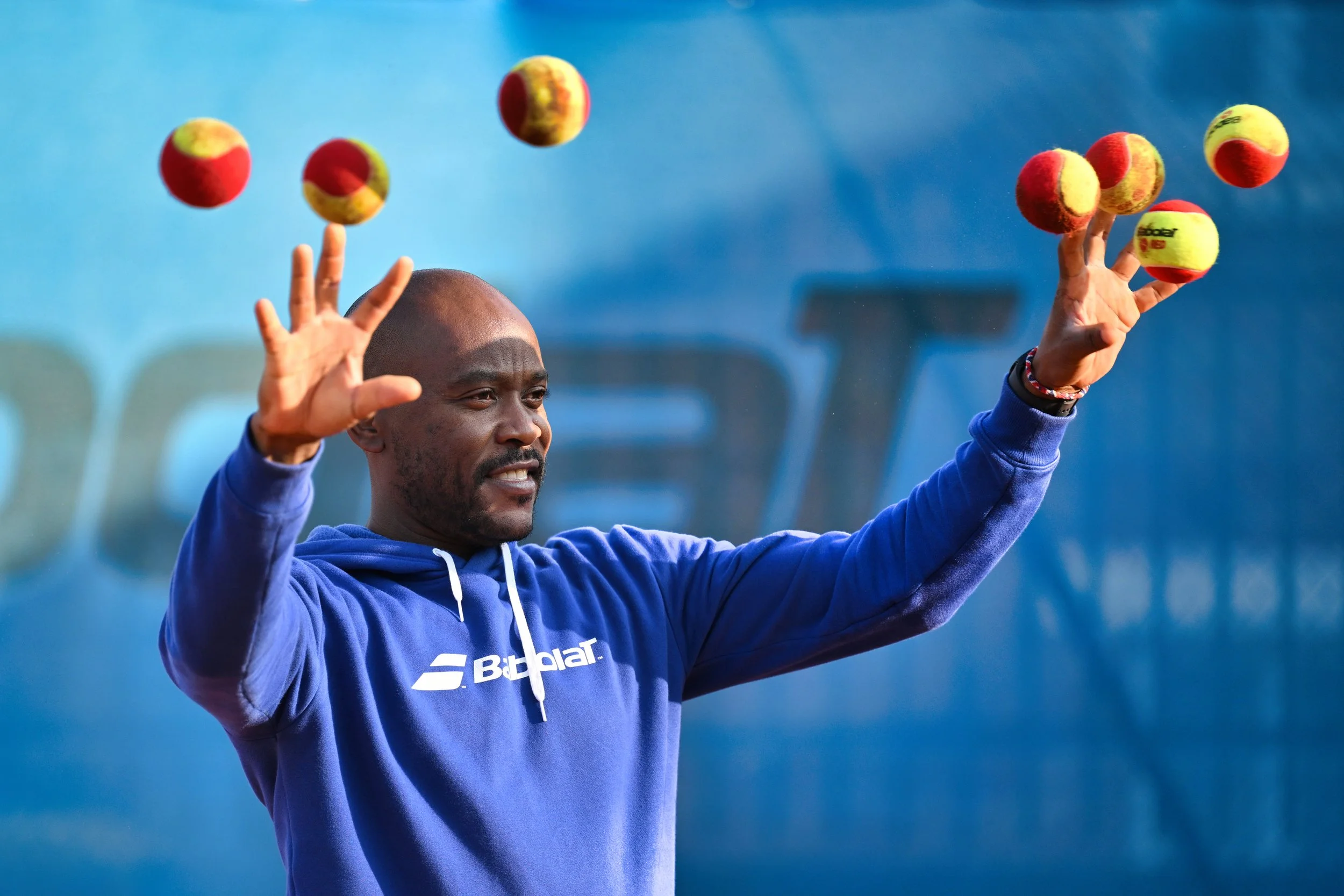 Un homme souriant en veste bleue, lançant des balles de tennis en l'air lors d'une séance d'entraînement ou d'un match sur un fond bleu.