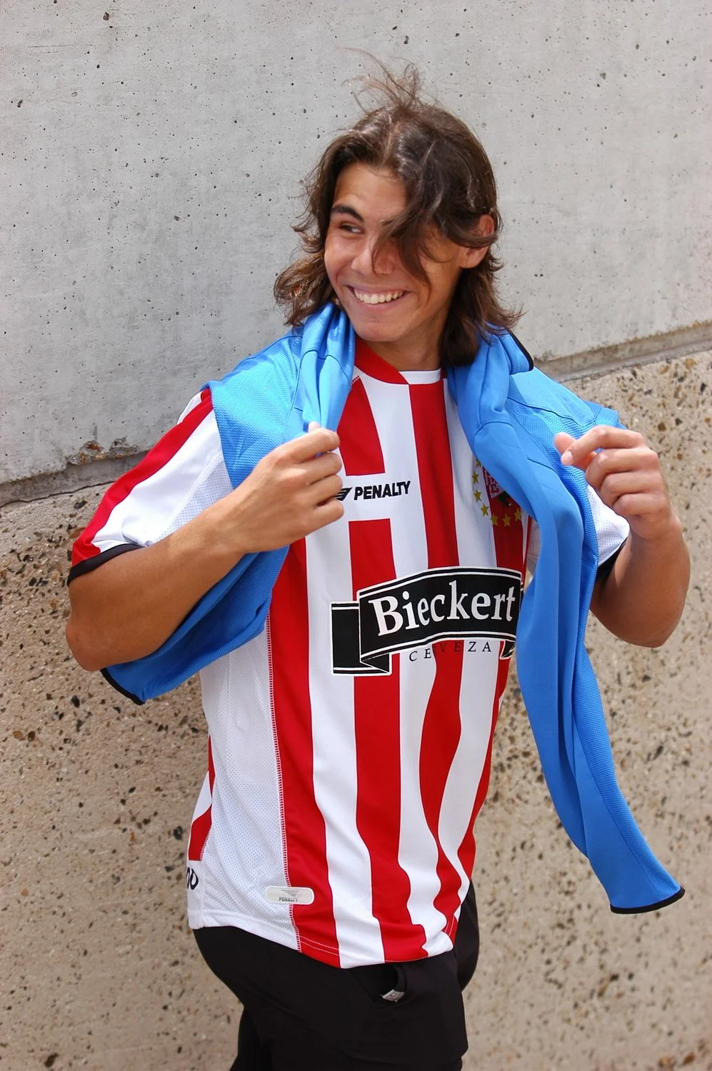Jeune homme souriant portant un maillot de football rouge et blanc rayé, avec un sweat bleu sur les épaules, devant un mur en béton.