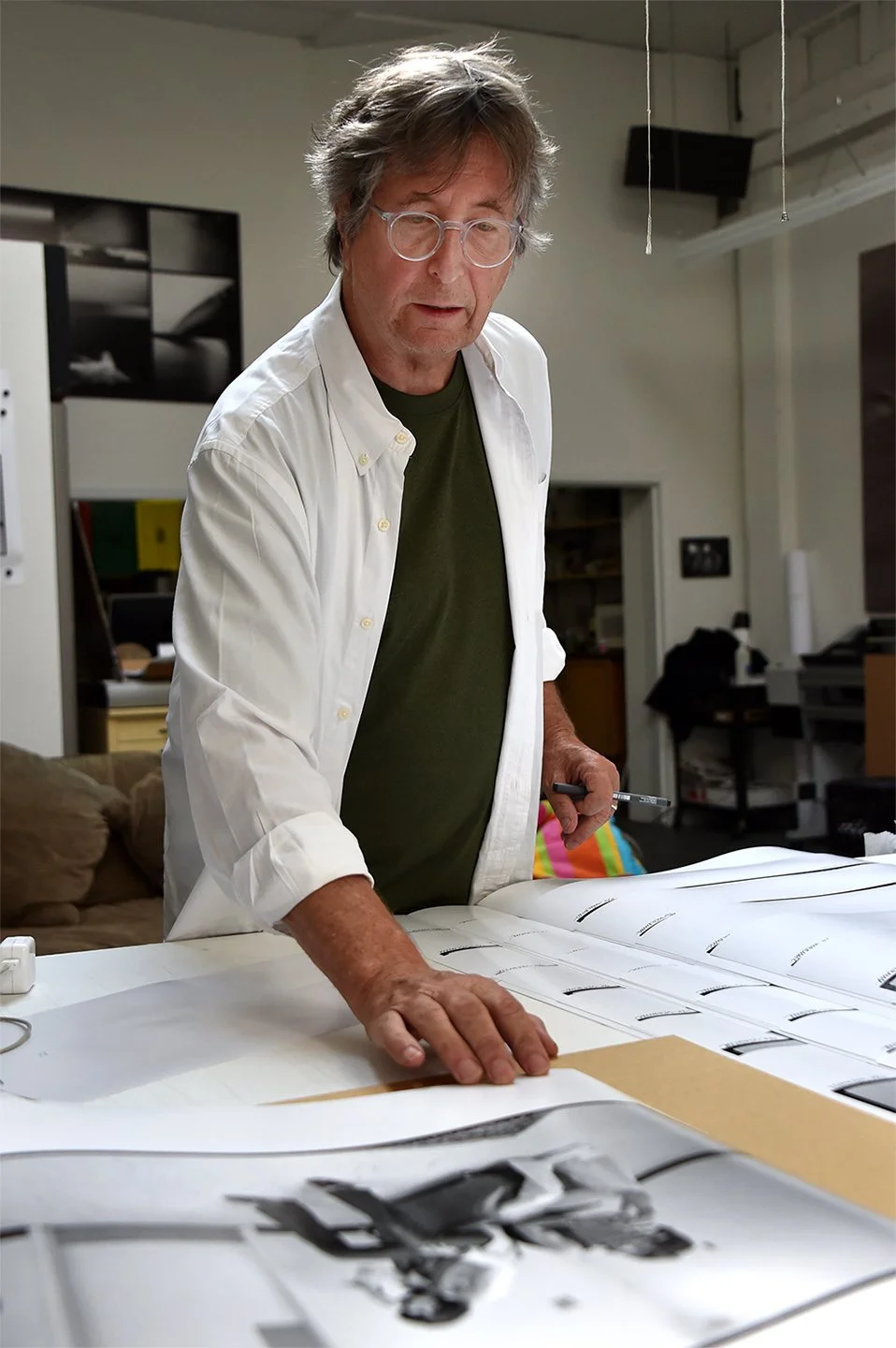 Un homme avec des lunettes, portant une chemise blanche par-dessus un t-shirt vert, examine des plans ou des dessins sur une grande table dans un espace intérieur.