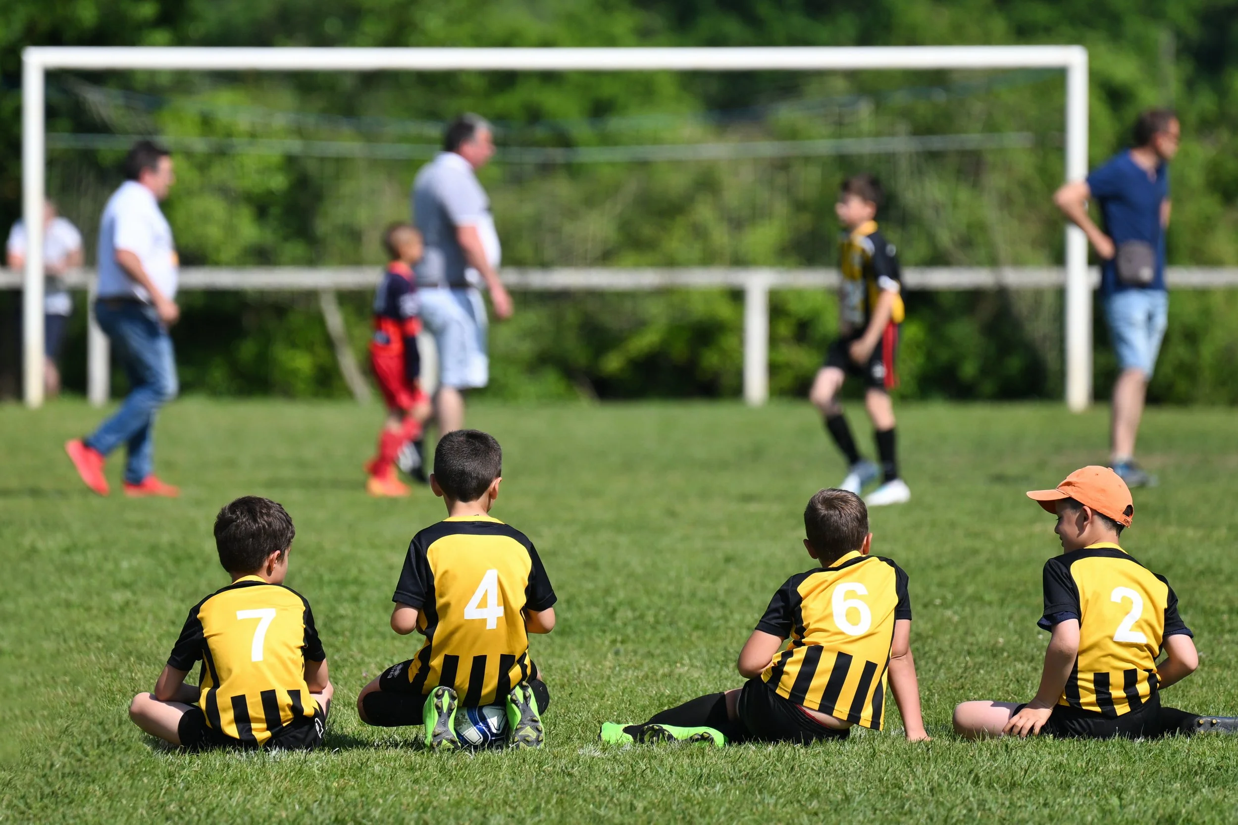 Jeunes joueurs de football en position assise sur l'herbe, portant des maillots jaunes avec des numéros, regardant un match sur un terrain en plein air. En arrière-plan, des enfants et des adultes regardent ou participent au jeu.