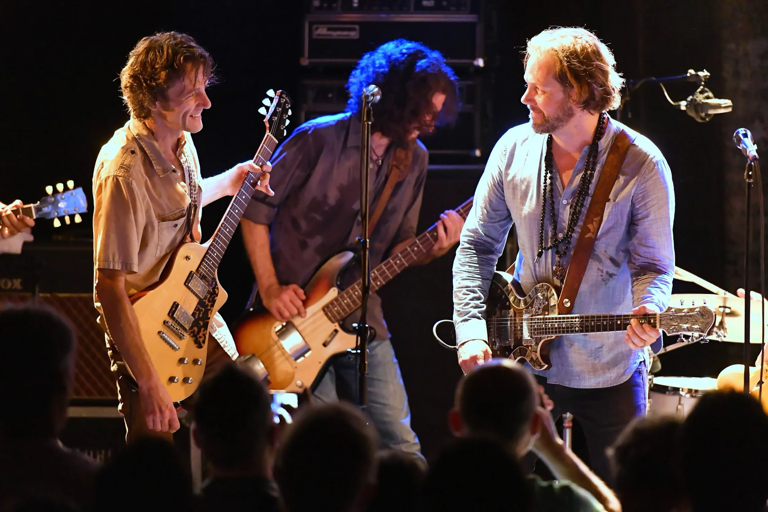 Groupe de musiciens jouant lors d'un concert, avec deux hommes à la guitare en avant, échangeant un sourire, et un autre musicien en arrière-plan.