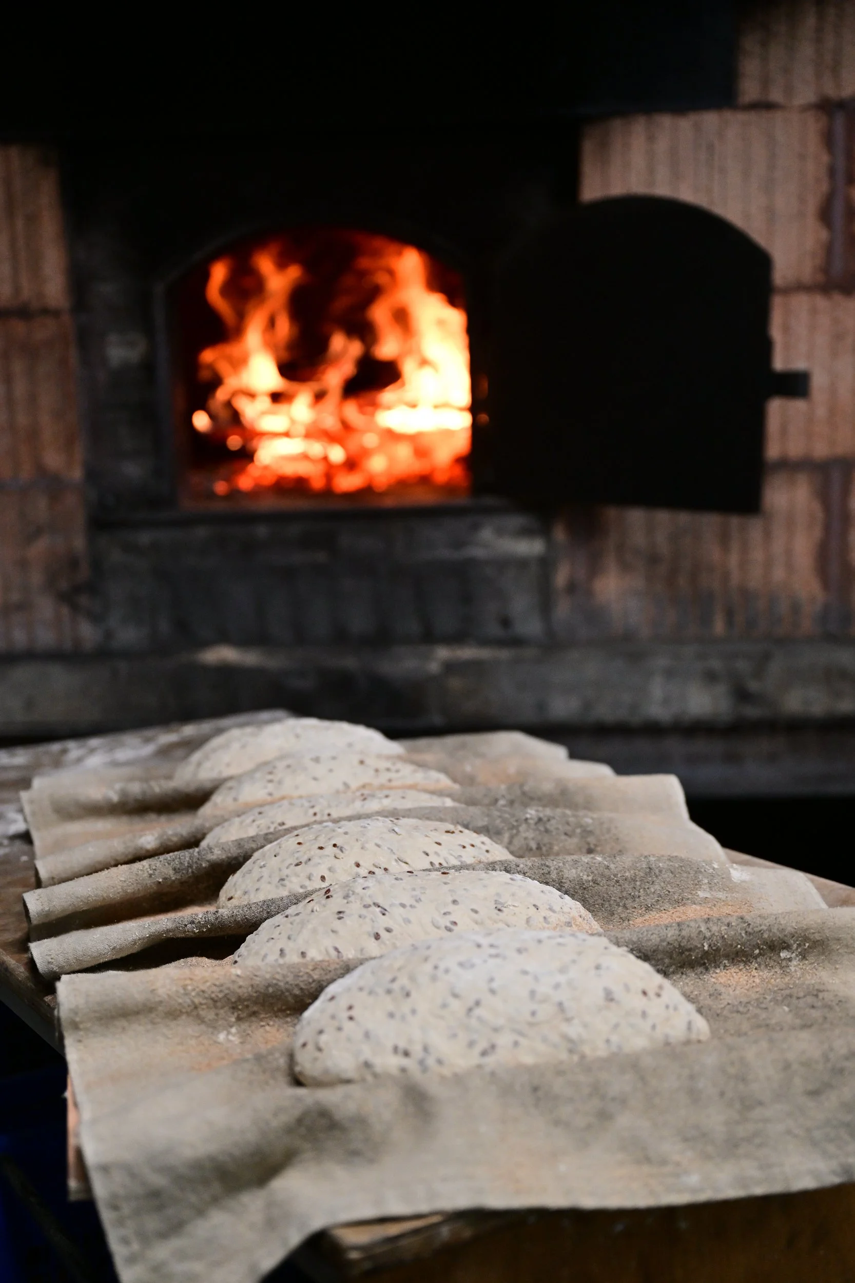 Un four à pizza en briques avec des pizzas non cuites garnies de graines et de fromage, prêtes à être enfournées.