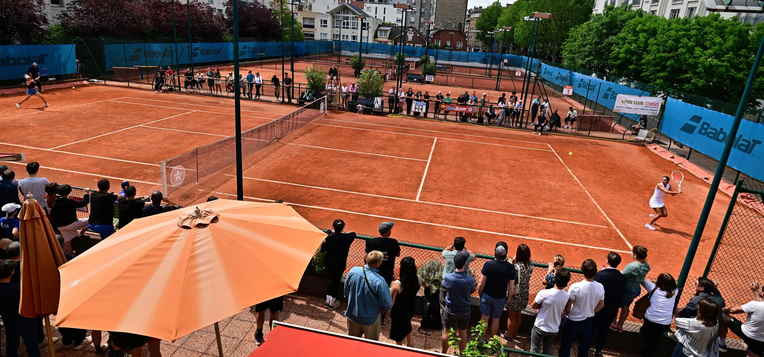 Une photo d'un court de tennis en terre battue avec des joueurs en action et un public qui regarde. Il y a des banderoles et des arbres verts en arrière-plan.