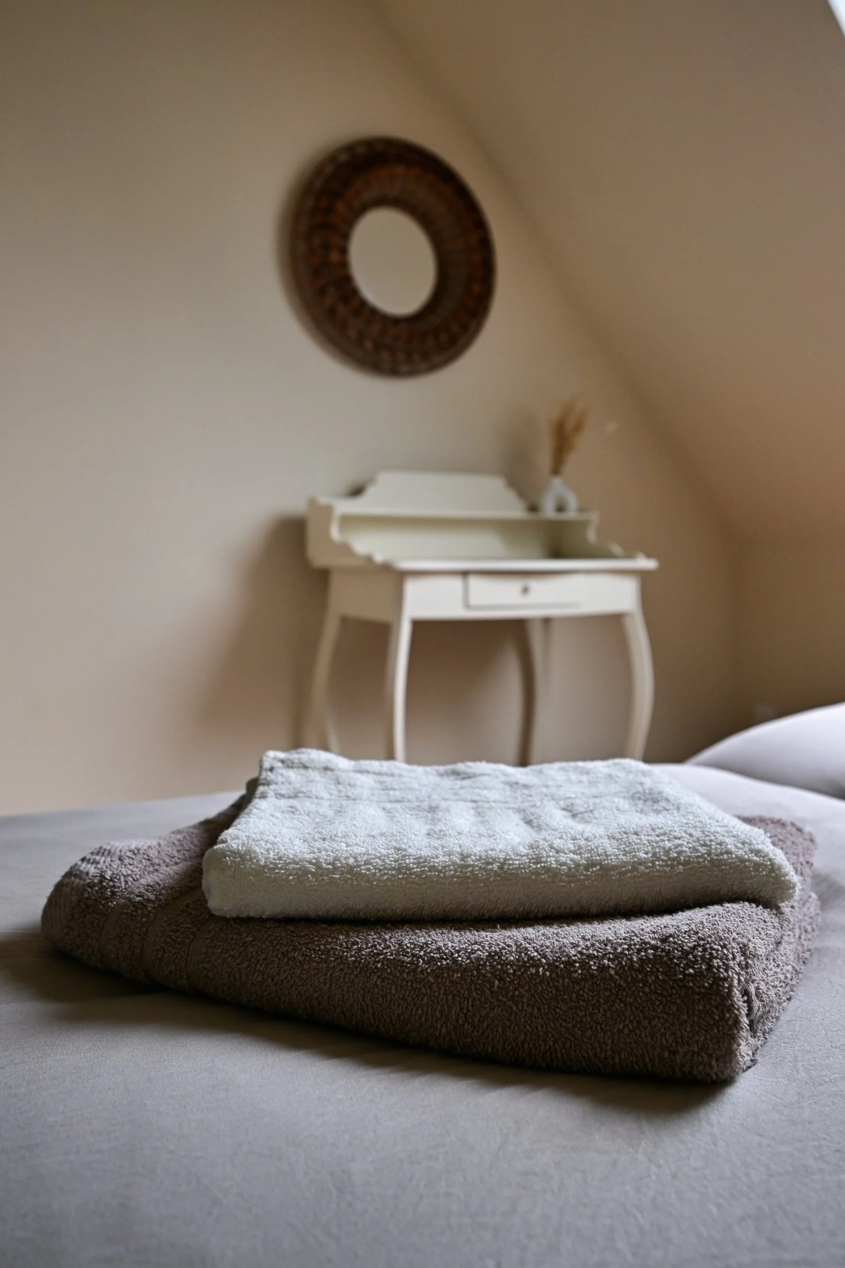 Une chambre avec une pile de deux serviettes pliées, une blanche en haut et une beige en dessous, posées sur un lit, avec un meuble blanc en arrière-plan, un miroir rond accroché au mur, et une décoration végétale dans un vase.