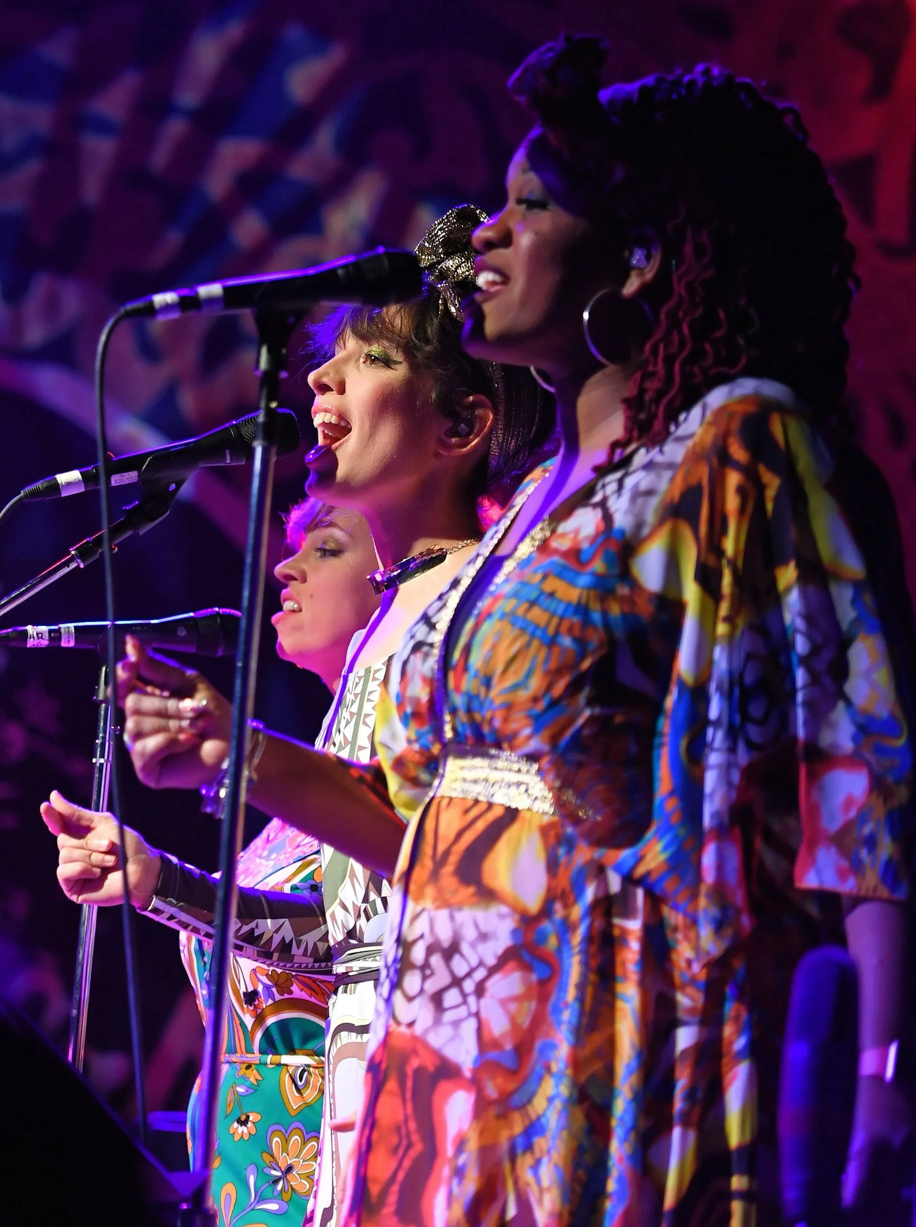 Trois femmes chantent dans un groupe, portant des vêtements colorés avec des motifs variés, sous un éclairage violet et rouge, devant un public.