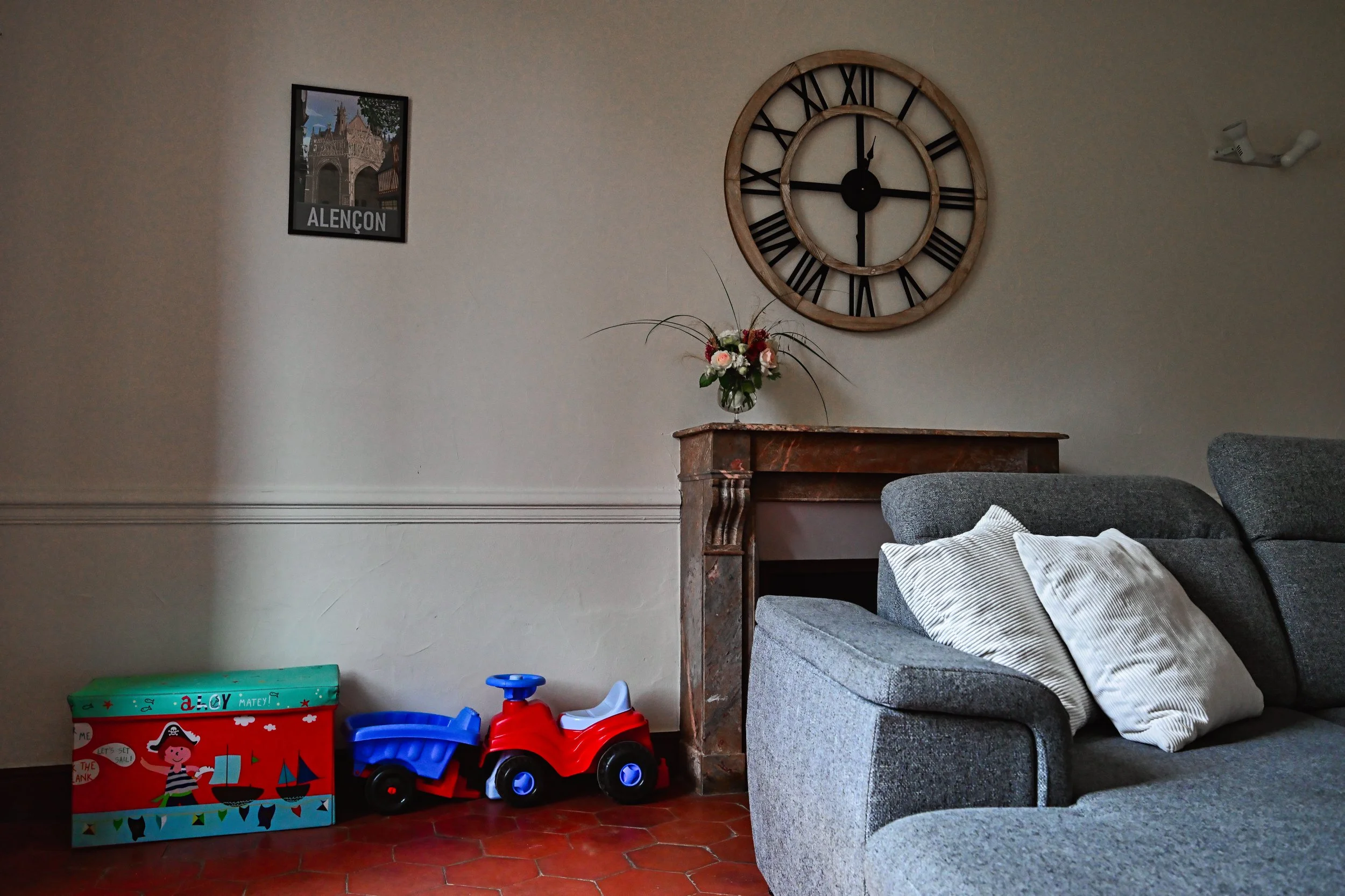 Salon avec un canapé gris, une table en bois, un vase avec des fleurs, une horloge murale en bois avec chiffres romains, une affiche d'Alençon, et des jouets d'enfants au sol.