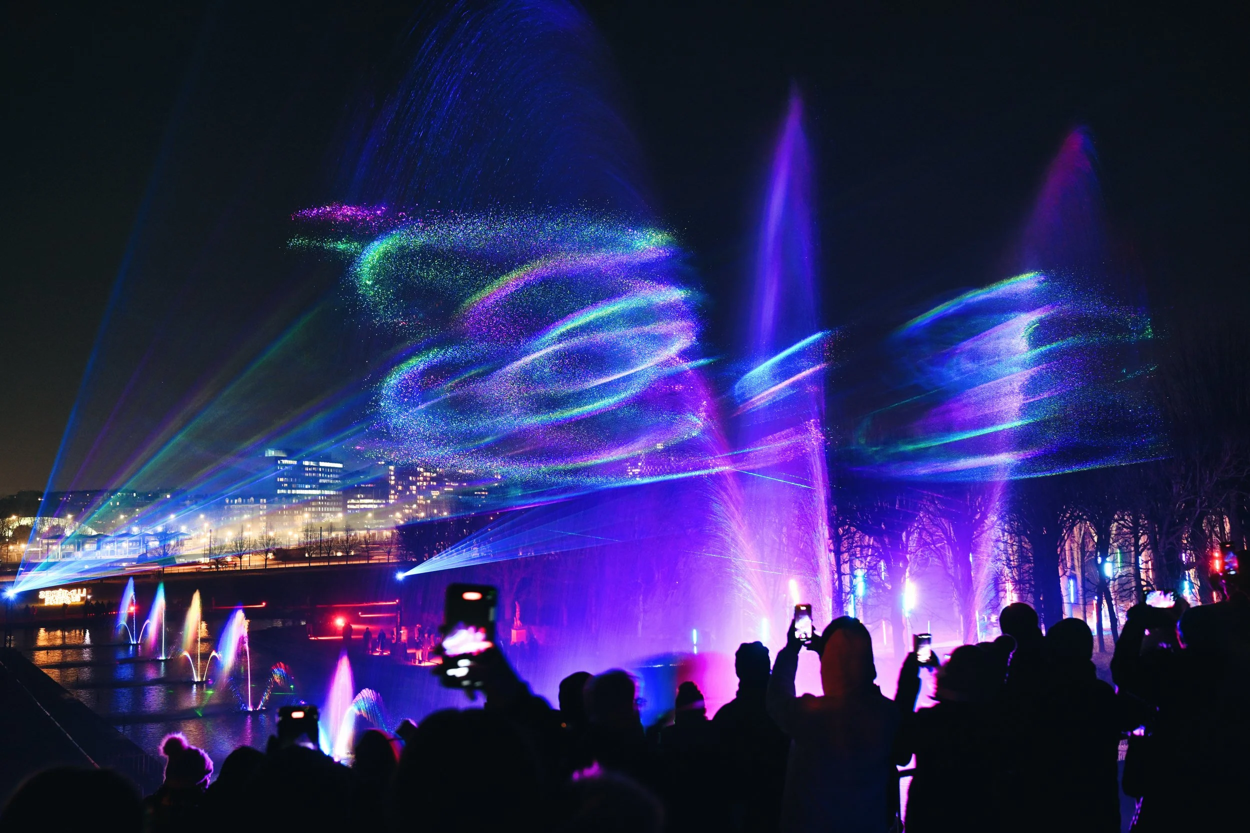Un spectacle nocturne avec des jets d'eau illuminés de couleurs violettes, bleues et vertes, avec une foule de spectateurs en silhouette en première scène.