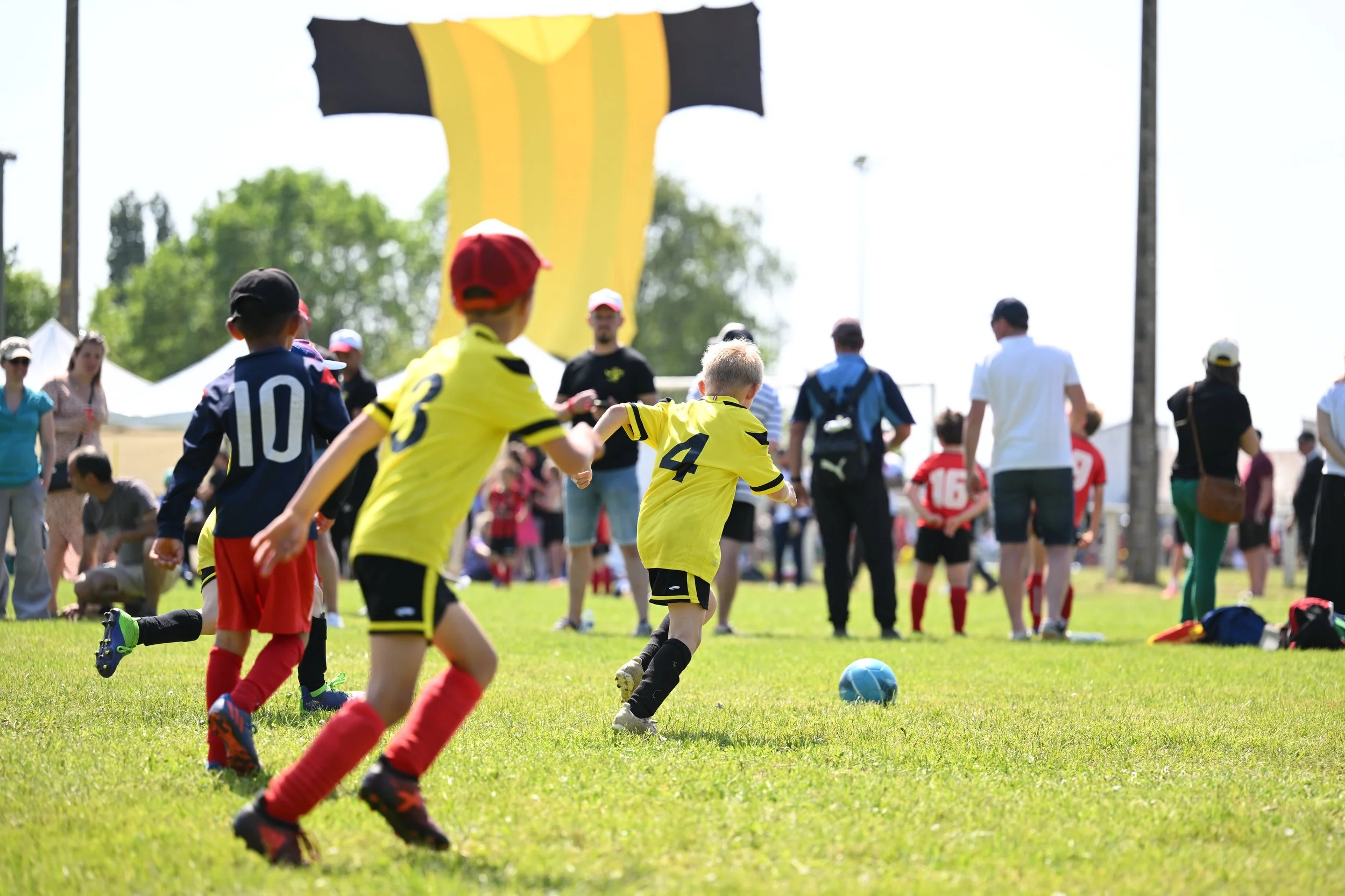 Jeunes enfants jouant au football lors d'un match en plein air avec des spectateurs autour, un ballon bleu sur l'herbe verte, un grand drapeau en arrière-plan et des gens qui regardent et prennent des photos.