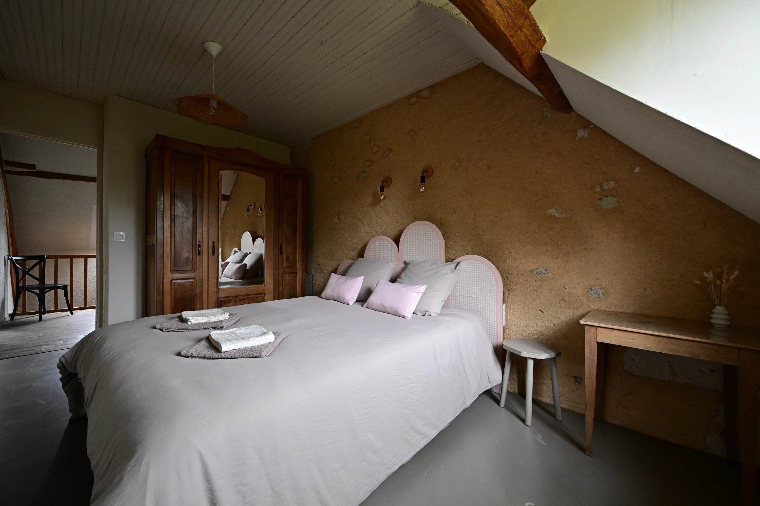 Chambre avec un lit double, deux coussins roses, une petite table, un miroir, une armoire en bois, et un mur en pierre avec des lampes murales.
