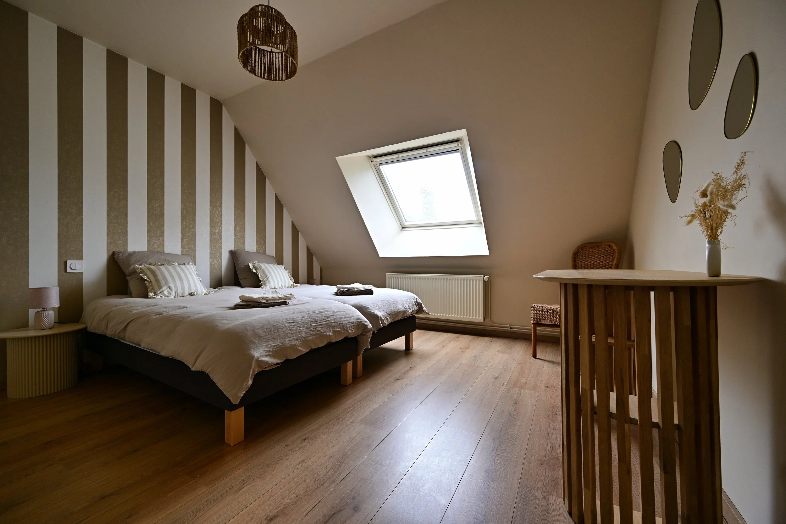 Chambre avec lit double, oreillers, table de chevet, lampes, plafond avec fenêtre de toit, décoration minimaliste, parquet en bois, mur à rayures verticales, mobilier en bois.