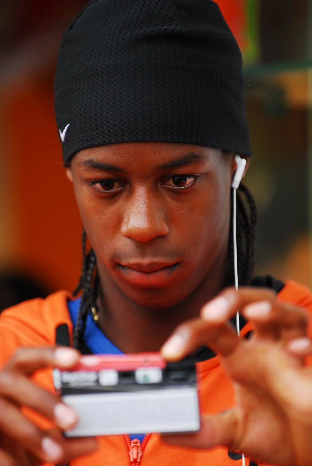 Jeune homme portant un bonnet noir, casque antibruit, et une veste orange, qui regarde son téléphone portable tout en portant des écouteurs.