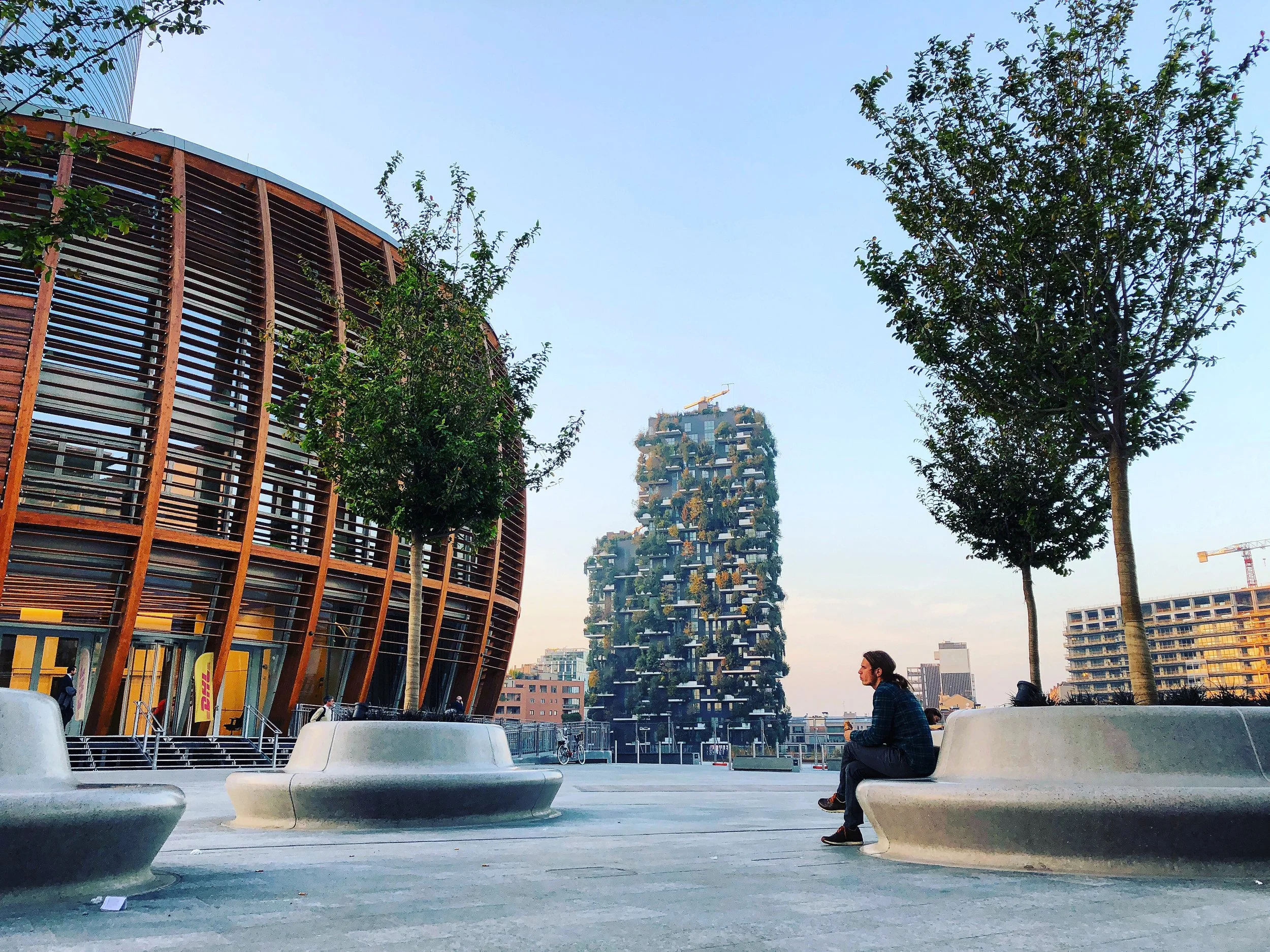 Une scène urbaine avec des bâtiments modernes, des arbres et une personne assise sur un banc, en fin d'après-midi ou en début de soirée.