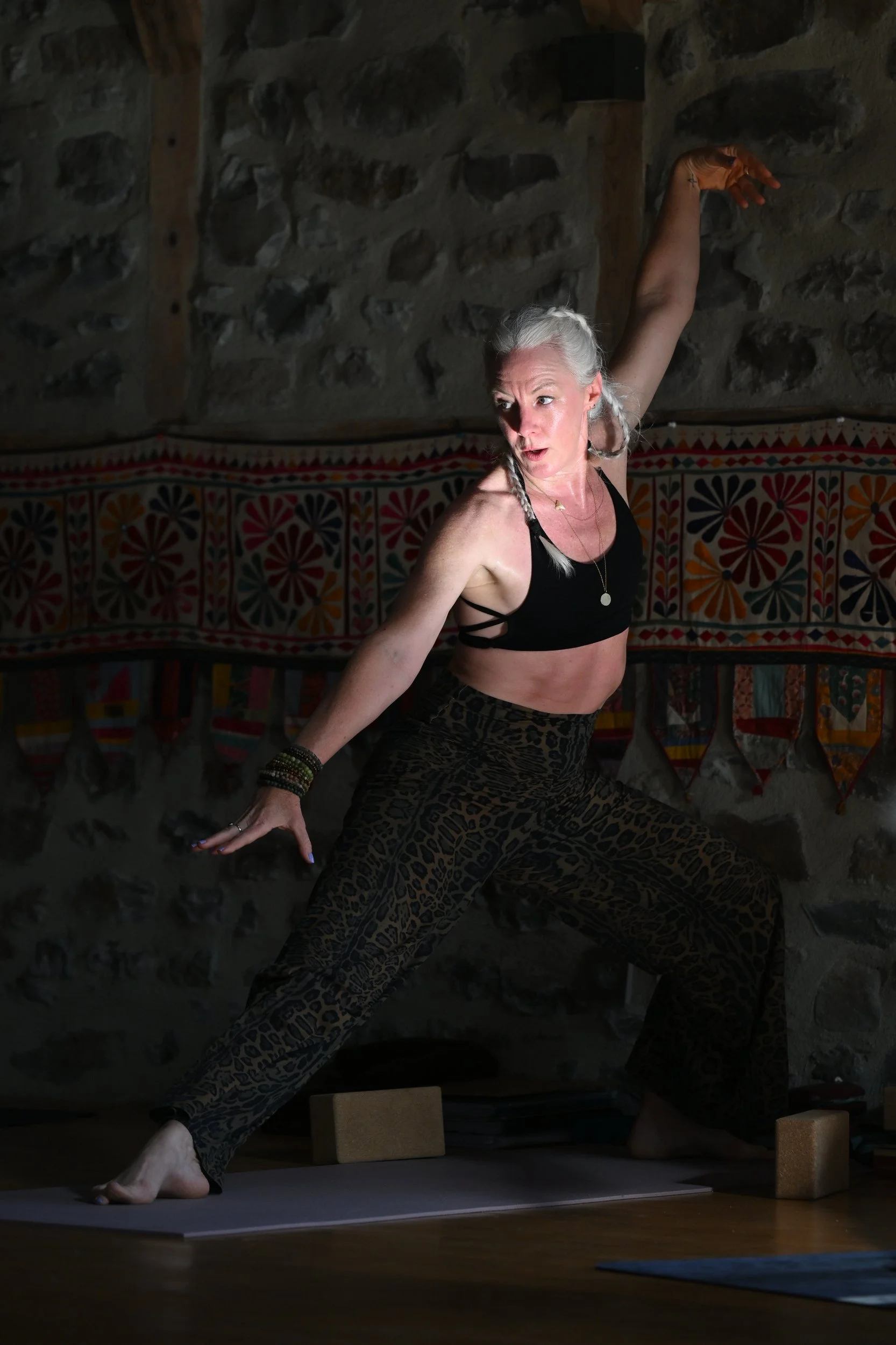 Femme pratiquant le yoga en position de guerrier dans une pièce à mur en pierre, portant un haut noir et un pantalon léopard, avec des blocs de yoga en dessous et un décor coloré en arrière-plan.