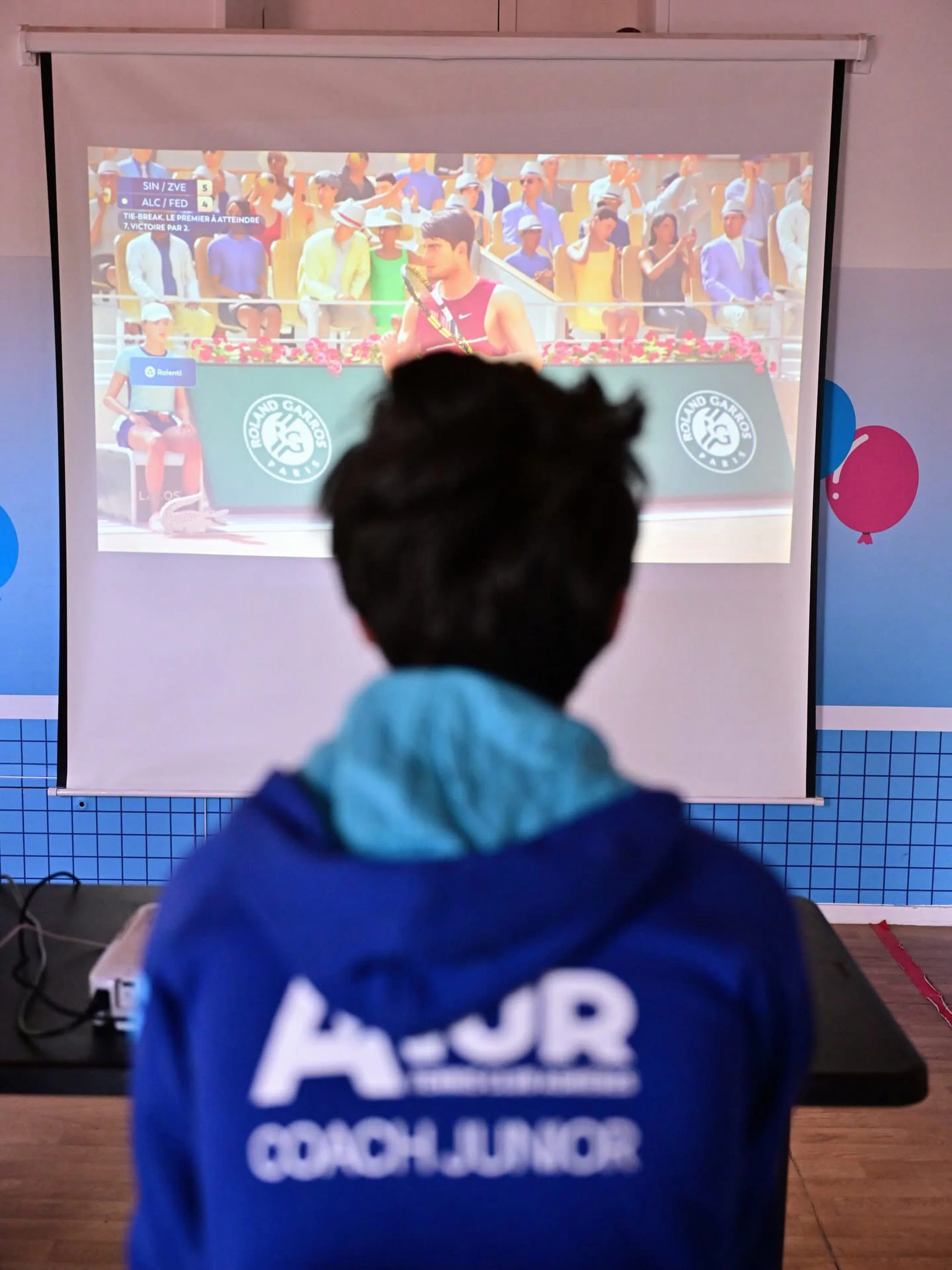 Un jeune homme regarde un match de tennis à travers une projection. Sur l'écran, on voit un joueur en pleine action avec un public nombreux en arrière-plan.