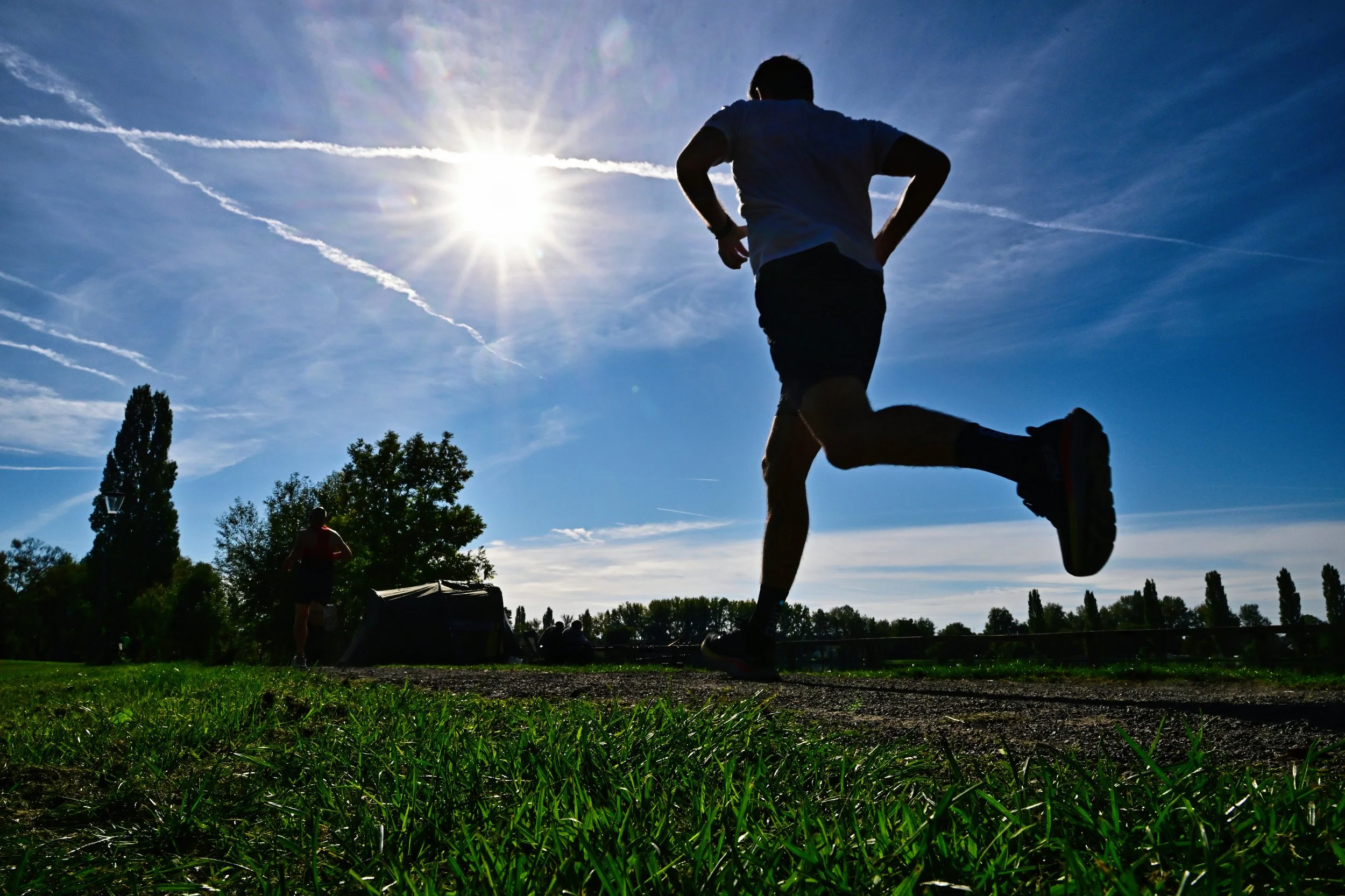 Une silhouette de personne en train de courir dans un parc sous un ciel ensoleillé, avec d'autres personnes au fond et des arbres.