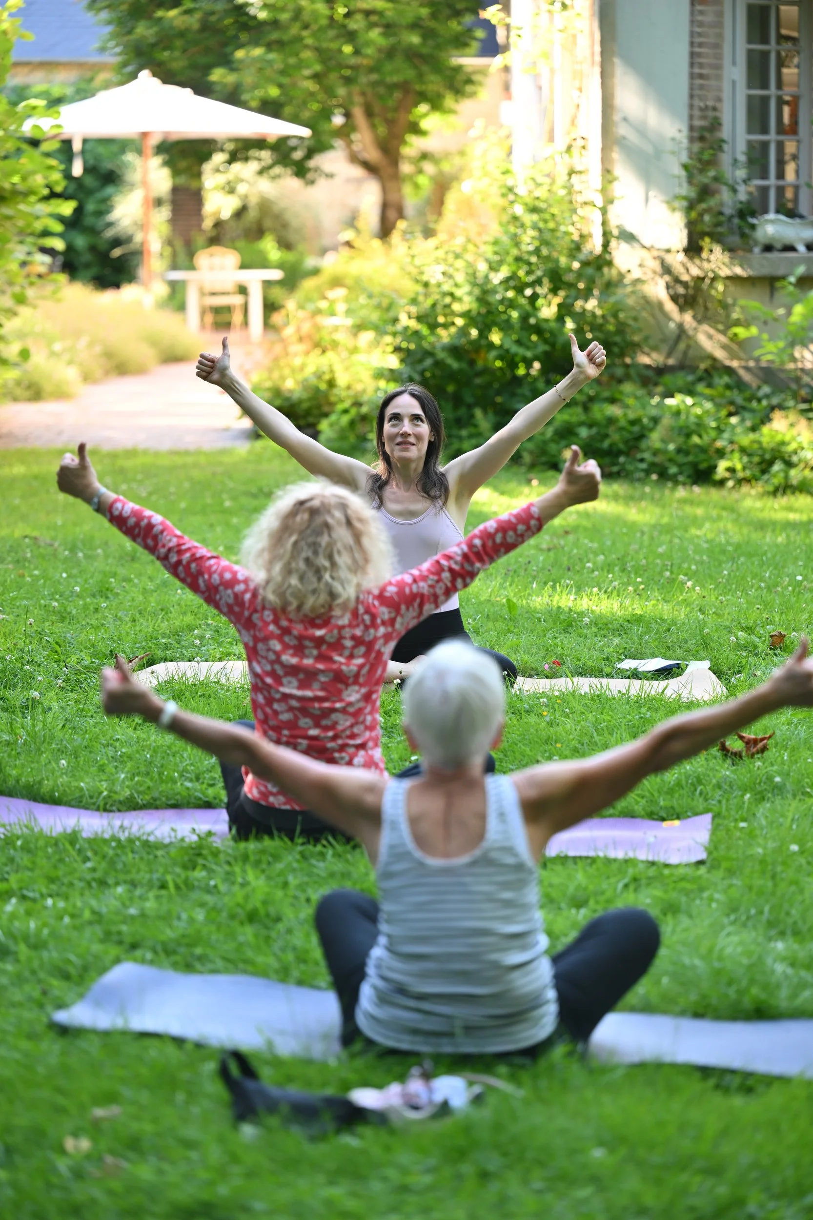 Groupe de personnes pratiquant le yoga ou la méditation dans un jardin verdoyant, en pleine nature, avec une femme en avant, les bras levés et un sourire.