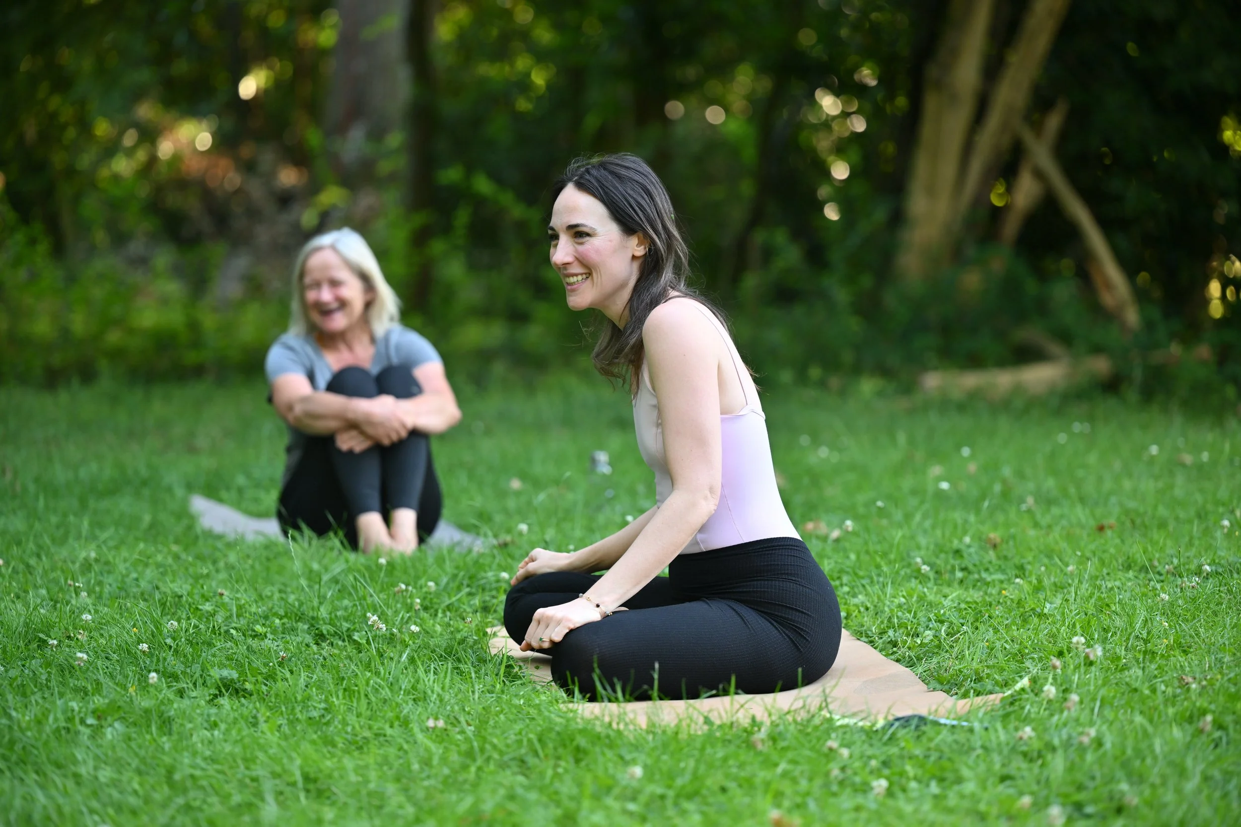 Deux femmes pratiquent le yoga dans un parc verdoyant, assises en position de lotus, en arrière-plan des arbres.