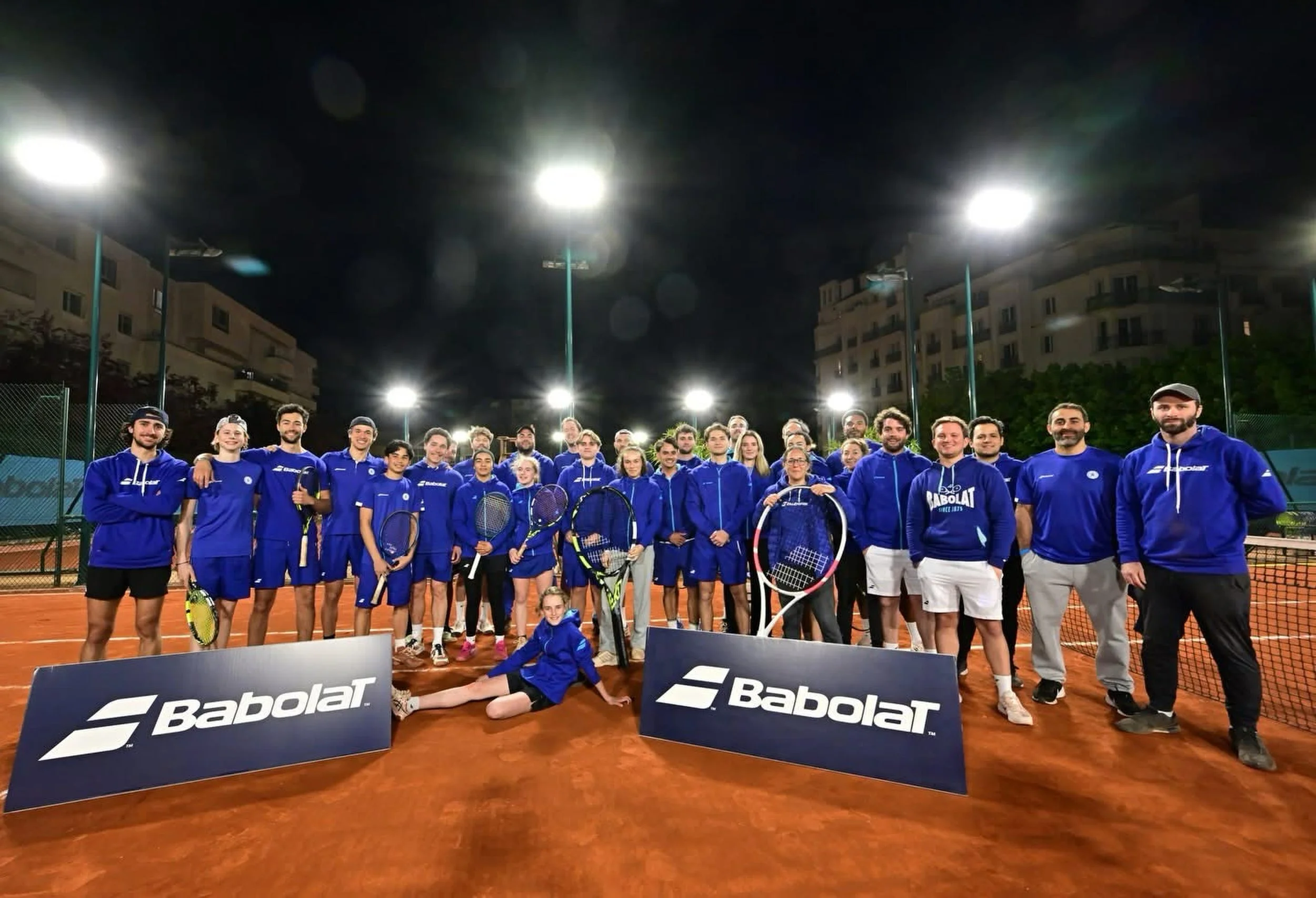 Groupe de personnes posant sur un terrain de tennis nocturne, portant des tenues de sport bleues, avec des raquettes, entourés de panneaux Babolat, sous un ciel noir avec plusieurs lampadaires lumineux.