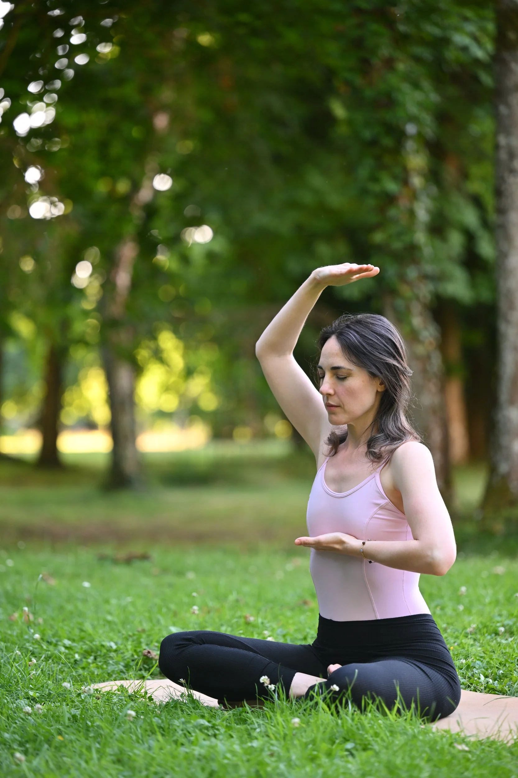 Une femme fait de la méditation en position de yoga dans un parc vert avec des arbres en arrière-plan, portant un haut rose et un pantalon noir.