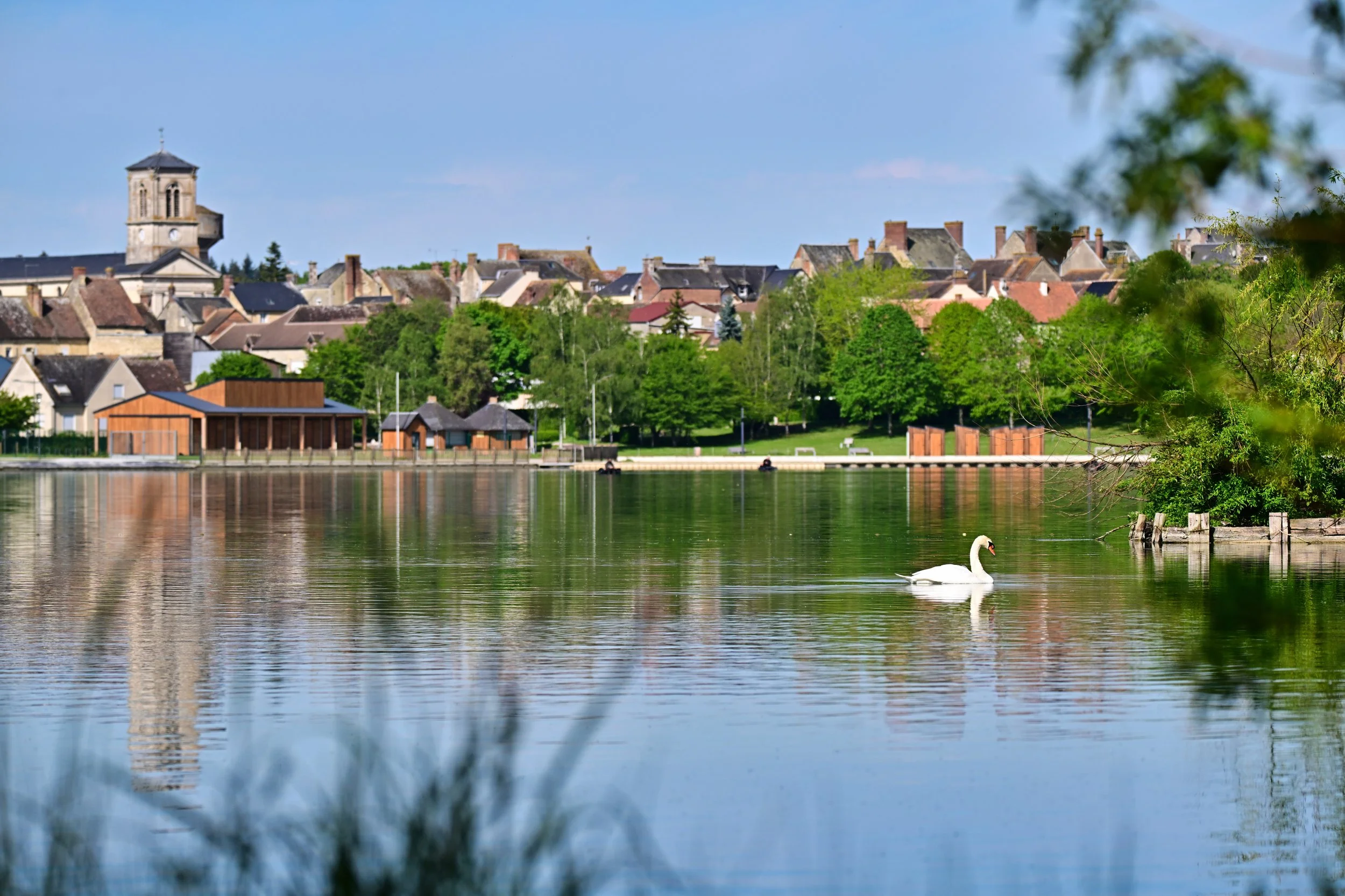 Une paisible vue d'un village avec des maisons en pierre, entouré d'arbres verts, vue depuis une rivière ou un lac avec un cygne nageant à la surface de l'eau.