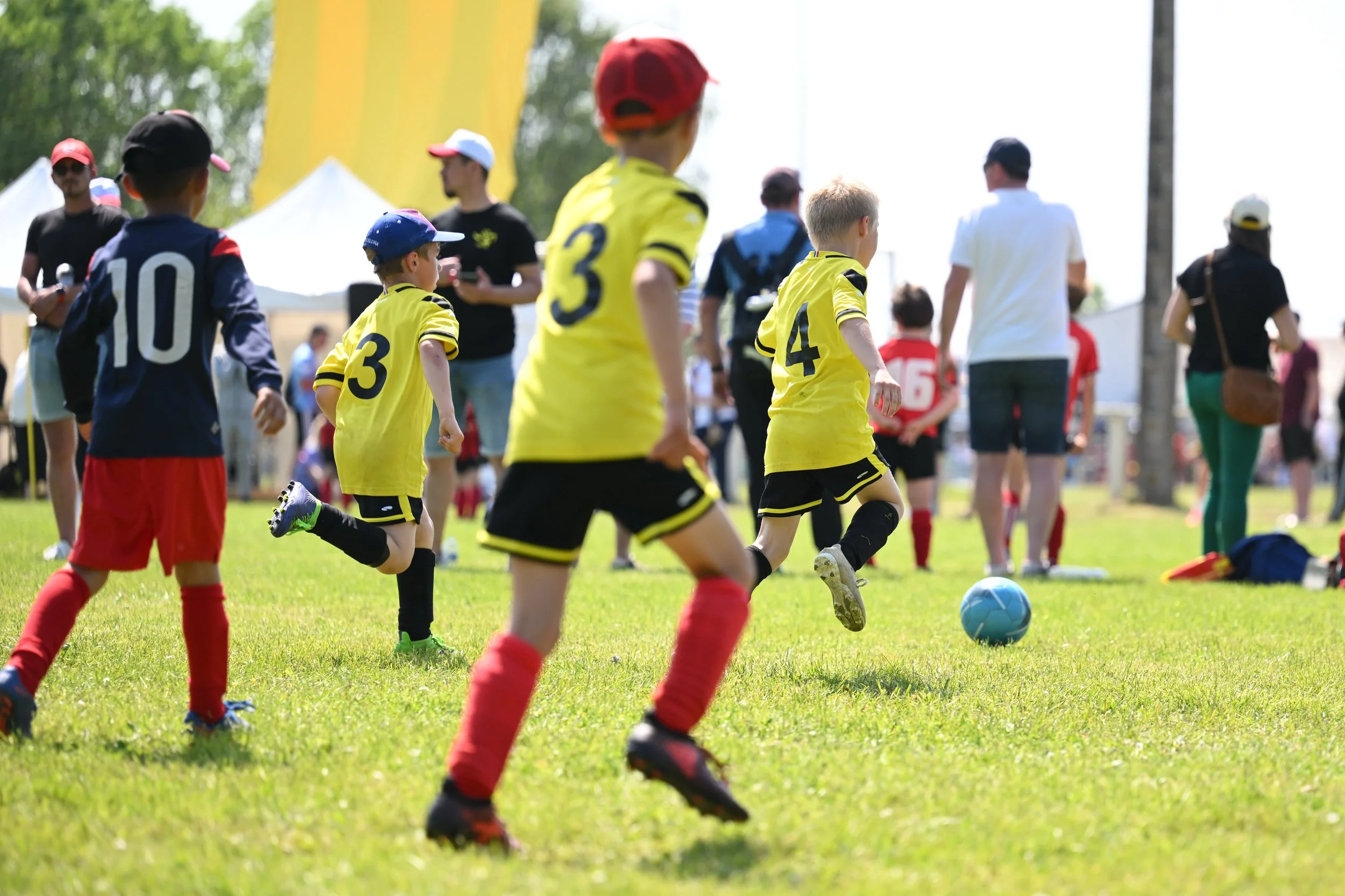 Jeunes enfants jouant au football sur un terrain en plein air, portant des maillots jaunes et noirs pour une équipe, et un joueur en maillot bleu et rouge. Il y a aussi des adultes en arrière-plan, probablement des entraîneurs ou des parents, dans un