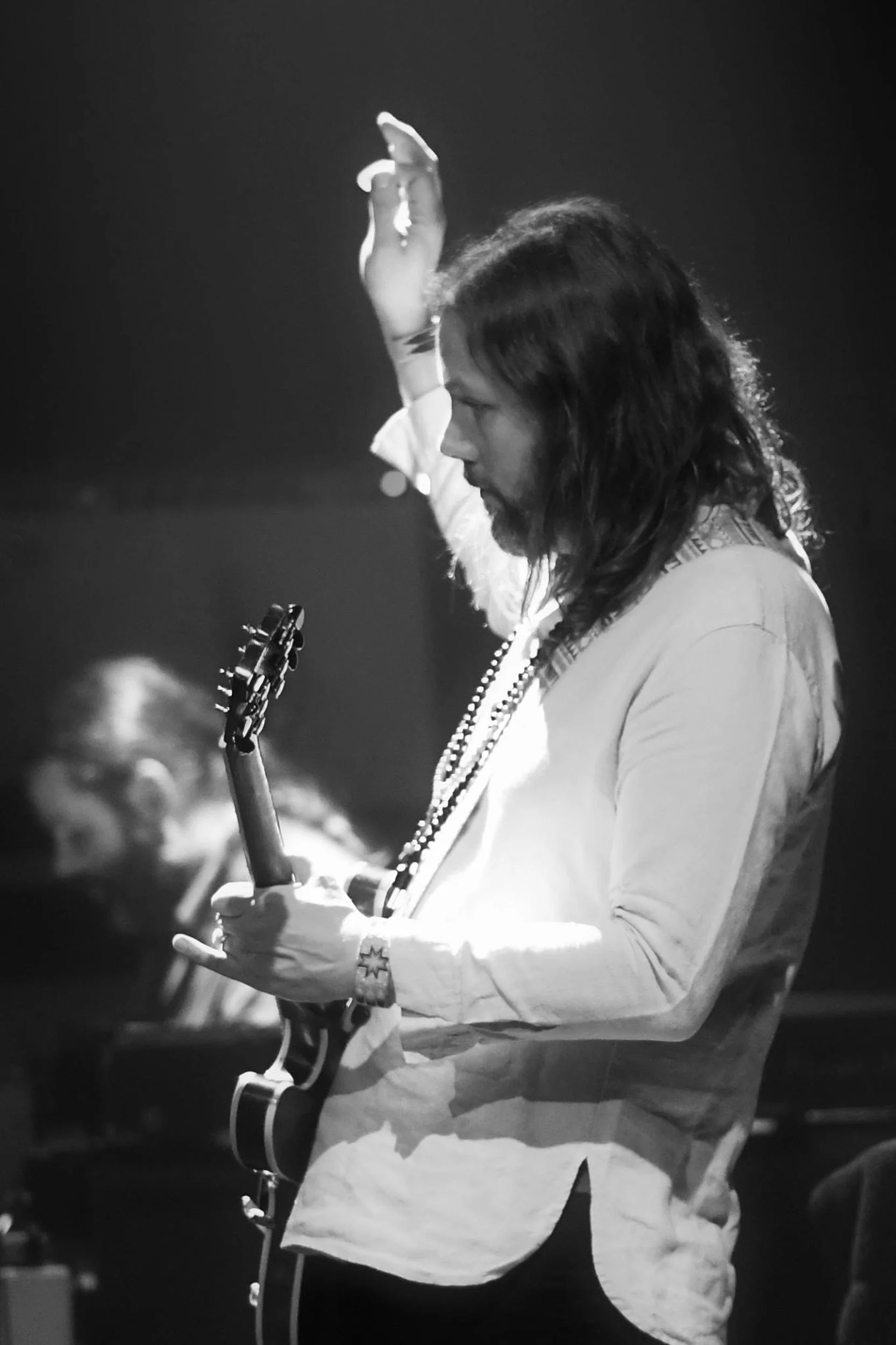 Un homme avec de longs cheveux noirs jouant de la guitare électrique, en noir et blanc, avec en arrière-plan d'autres musiciens ou personnes, lors d'une performance ou d'un concert.