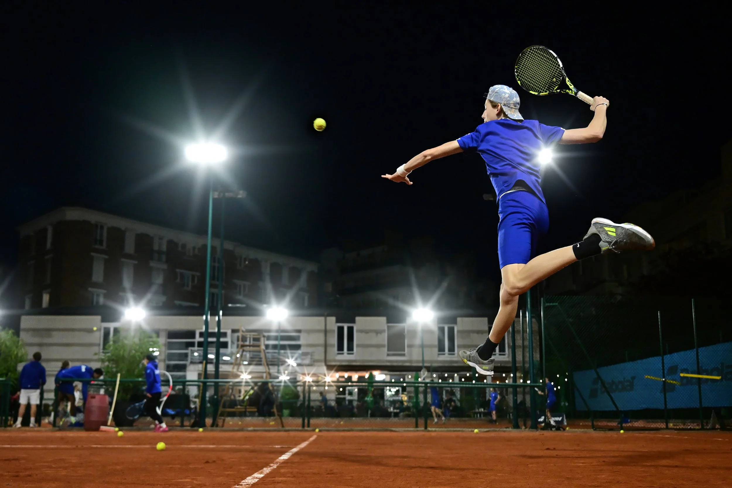 Un jeune joueur de tennis effectue un smash lors d'un match nocturne sous des projecteurs, avec d'autres personnes et des bâtiments en arrière-plan.