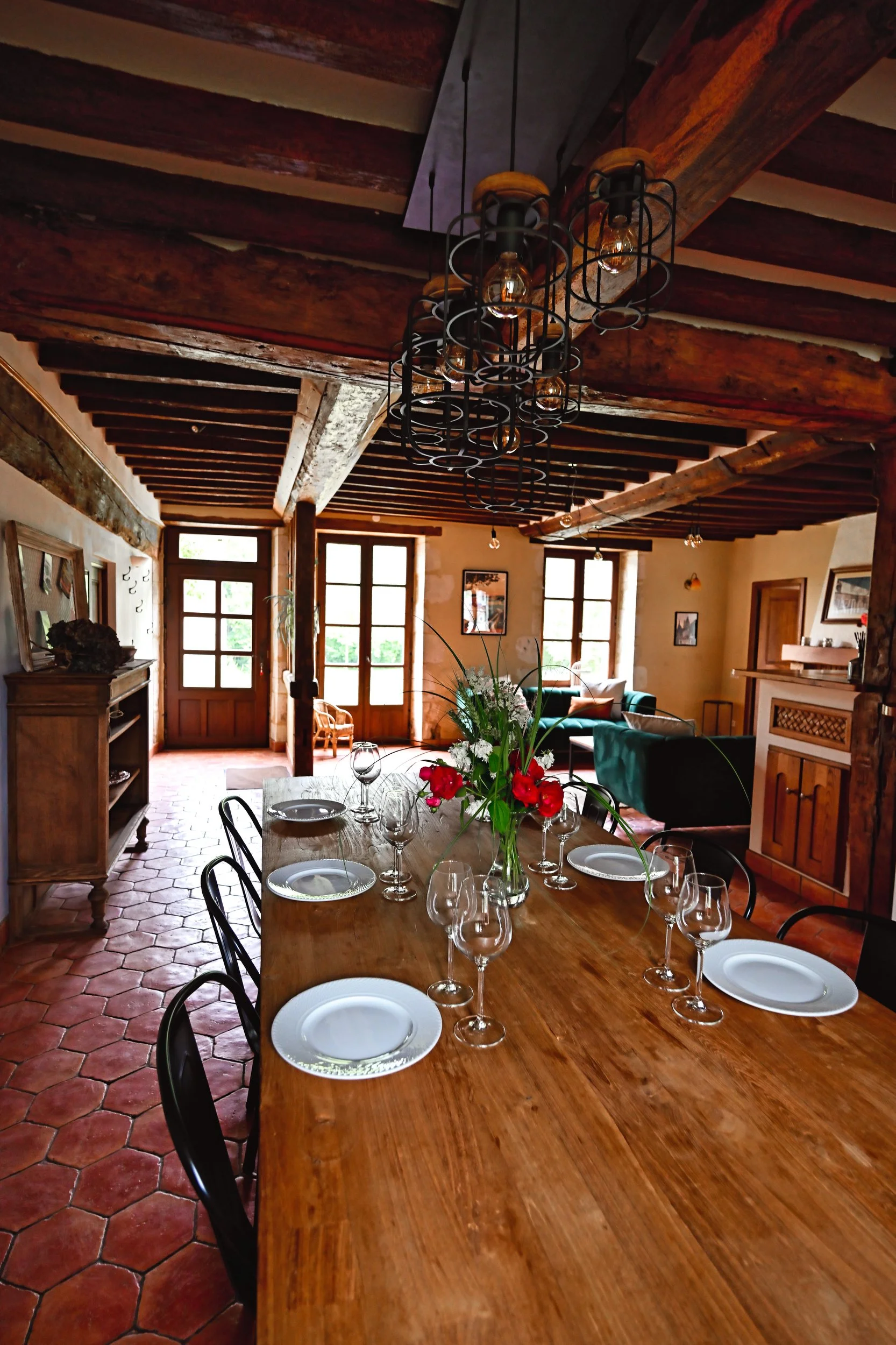 Intérieur d'une salle à manger chaleureuse avec un plafond en bois, une grande table en bois, des assiettes, des verres à vin, un bouquet de fleurs rouges sur la table, et des fenêtres laissant entrer la lumière naturelle.