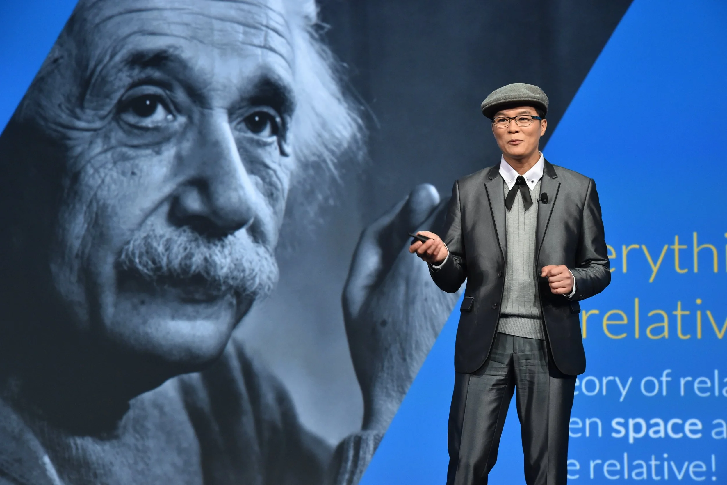 Un homme parle lors d'une présentation devant une grande image en noir et blanc d'Albert Einstein, avec un fond bleu incluant du texte.